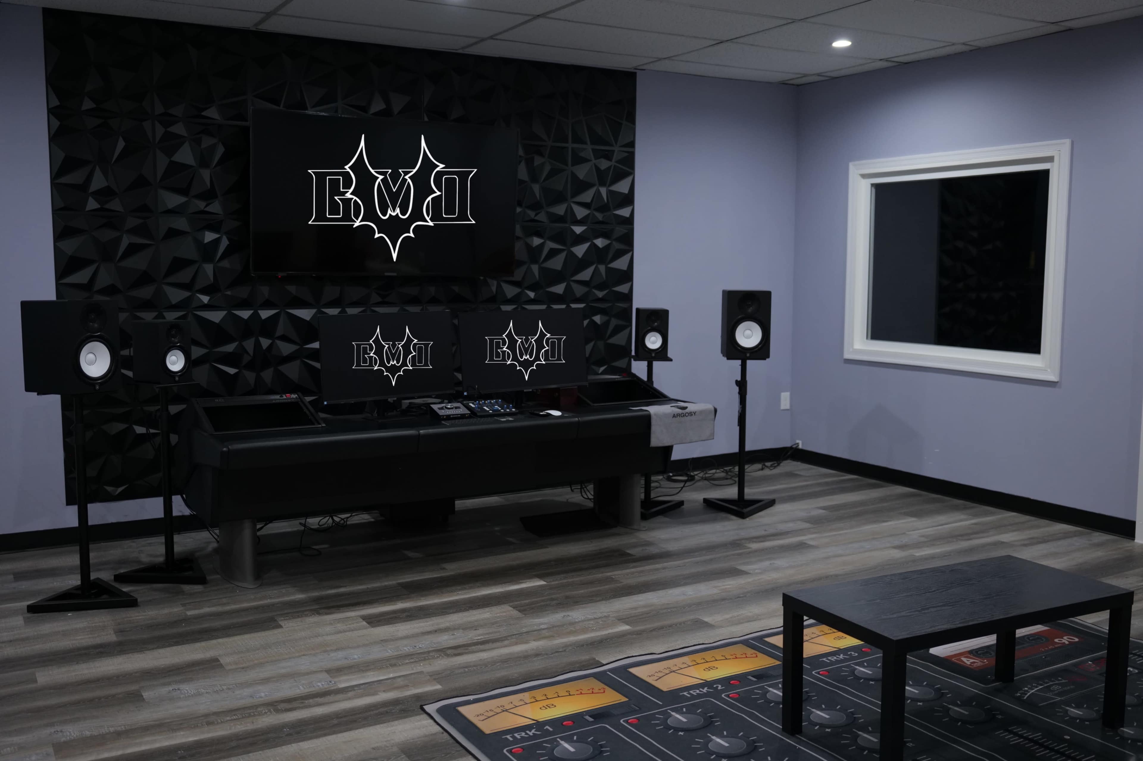 A modern music studio with dark acoustic paneling, a central mixing console, speakers on either side, and a large monitor displaying a logo on the wall.