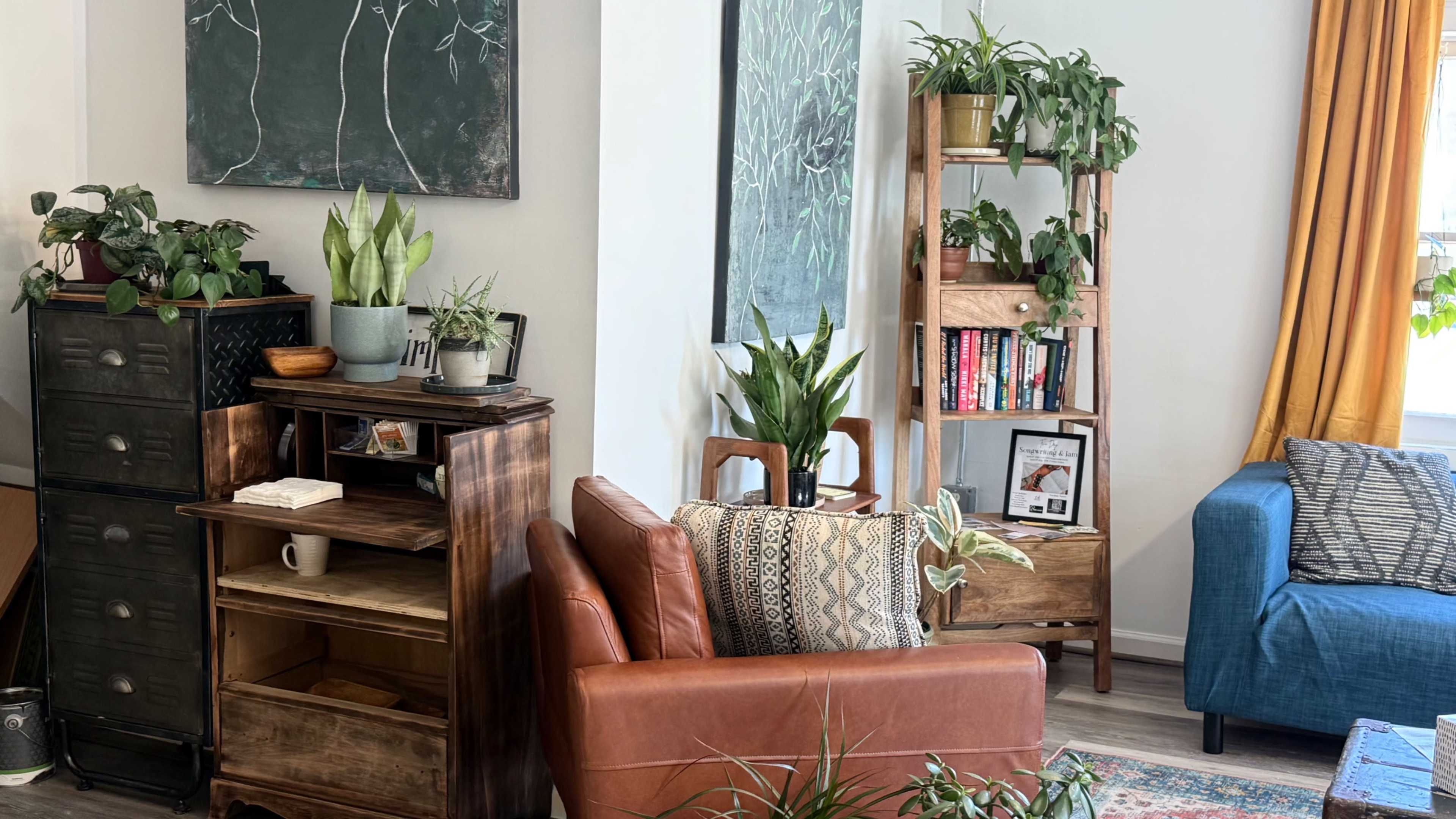 The image depicts a cozy living room with a leather armchair, a wooden bookshelf filled with books, and various potted plants arranged throughout the space.