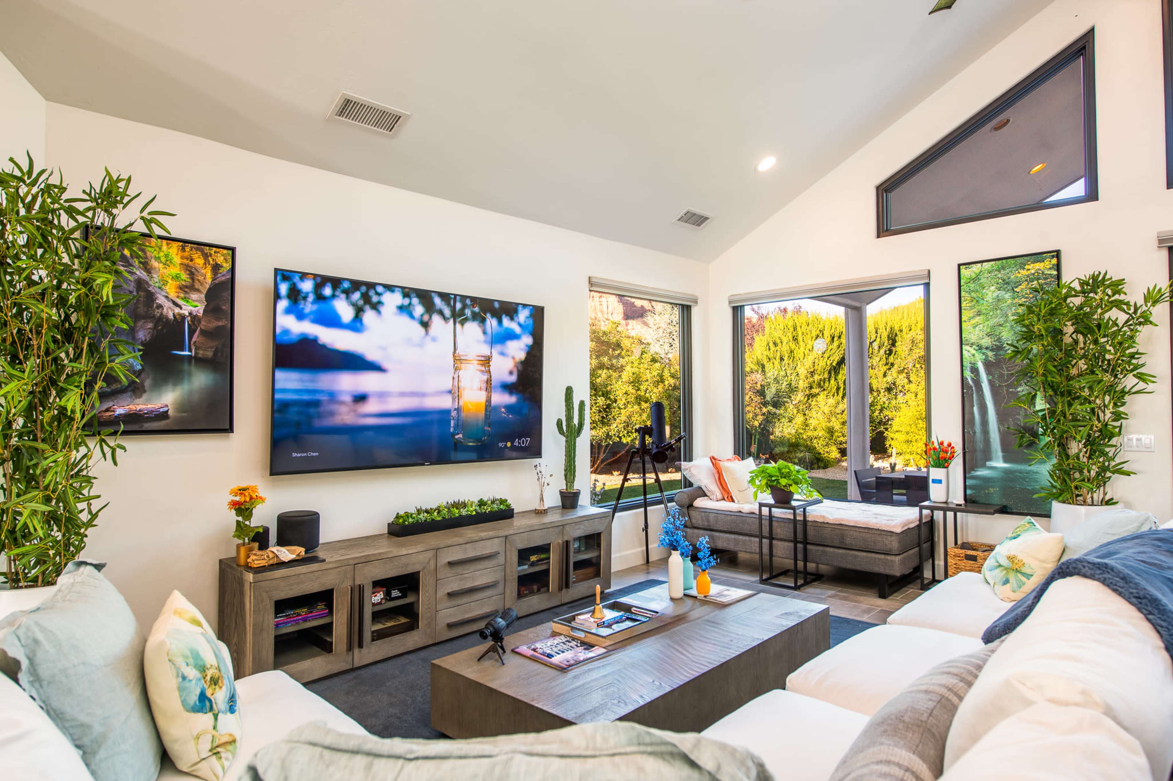 A modern living room with a large television, a coffee table, and windows overlooking a garden, all surrounded by potted plants.