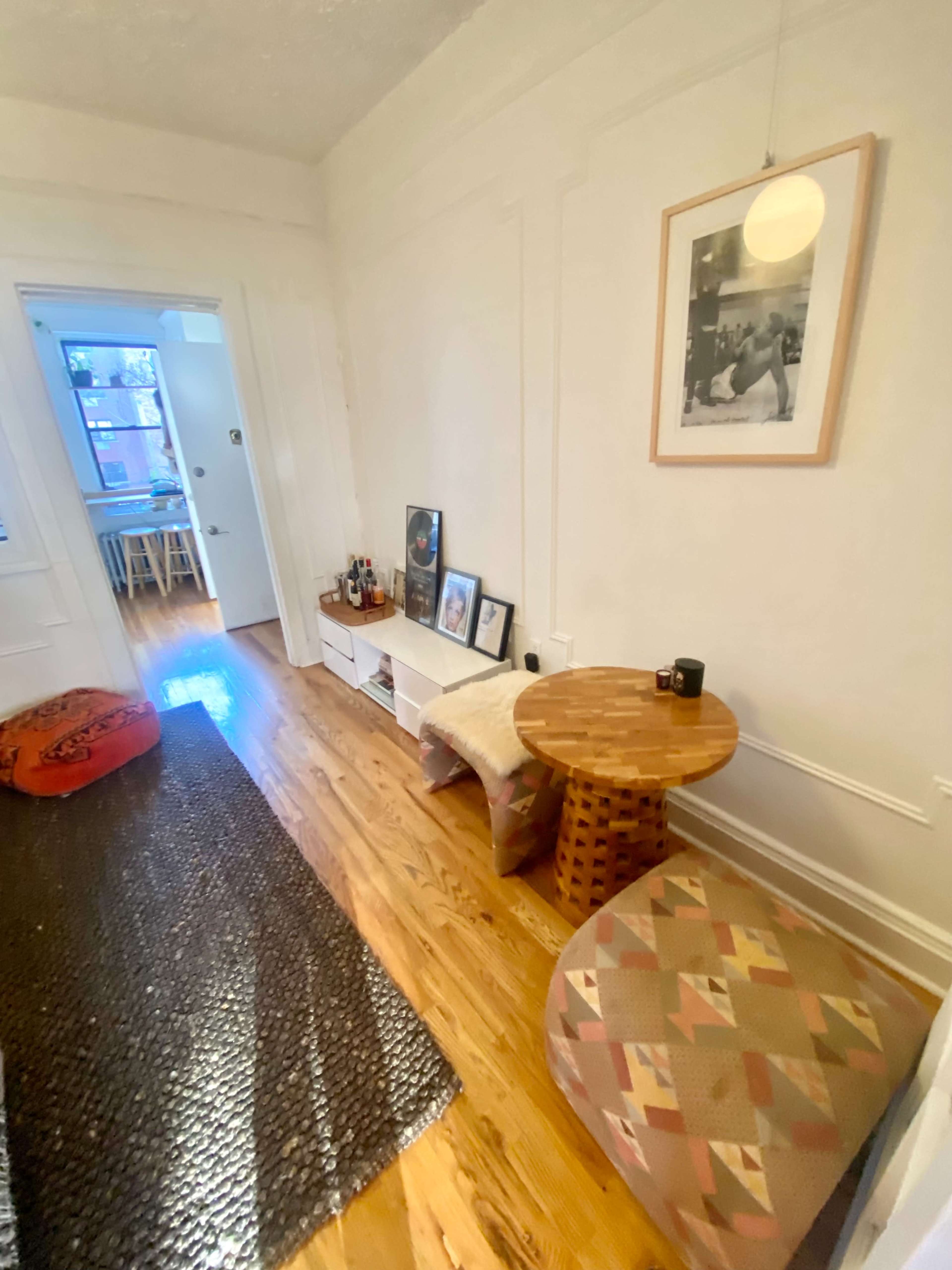 The image shows a narrow hallway with wooden flooring, a woven rug, a small shelf displaying framed photos and bottles, and a couple of patterned seating cushions.