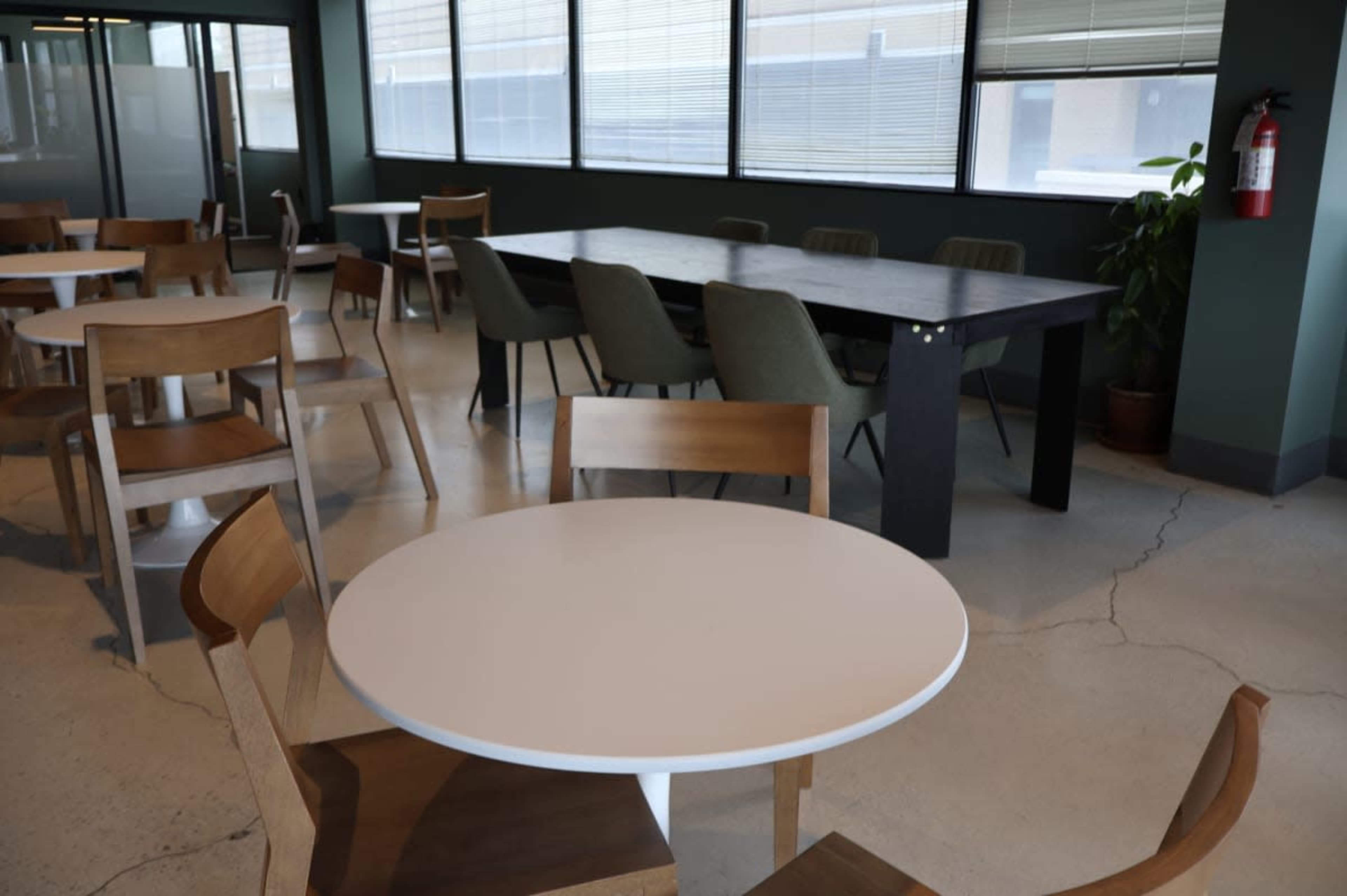 The image shows a modern dining space with several tables and chairs, featuring large windows and a plant in the corner.