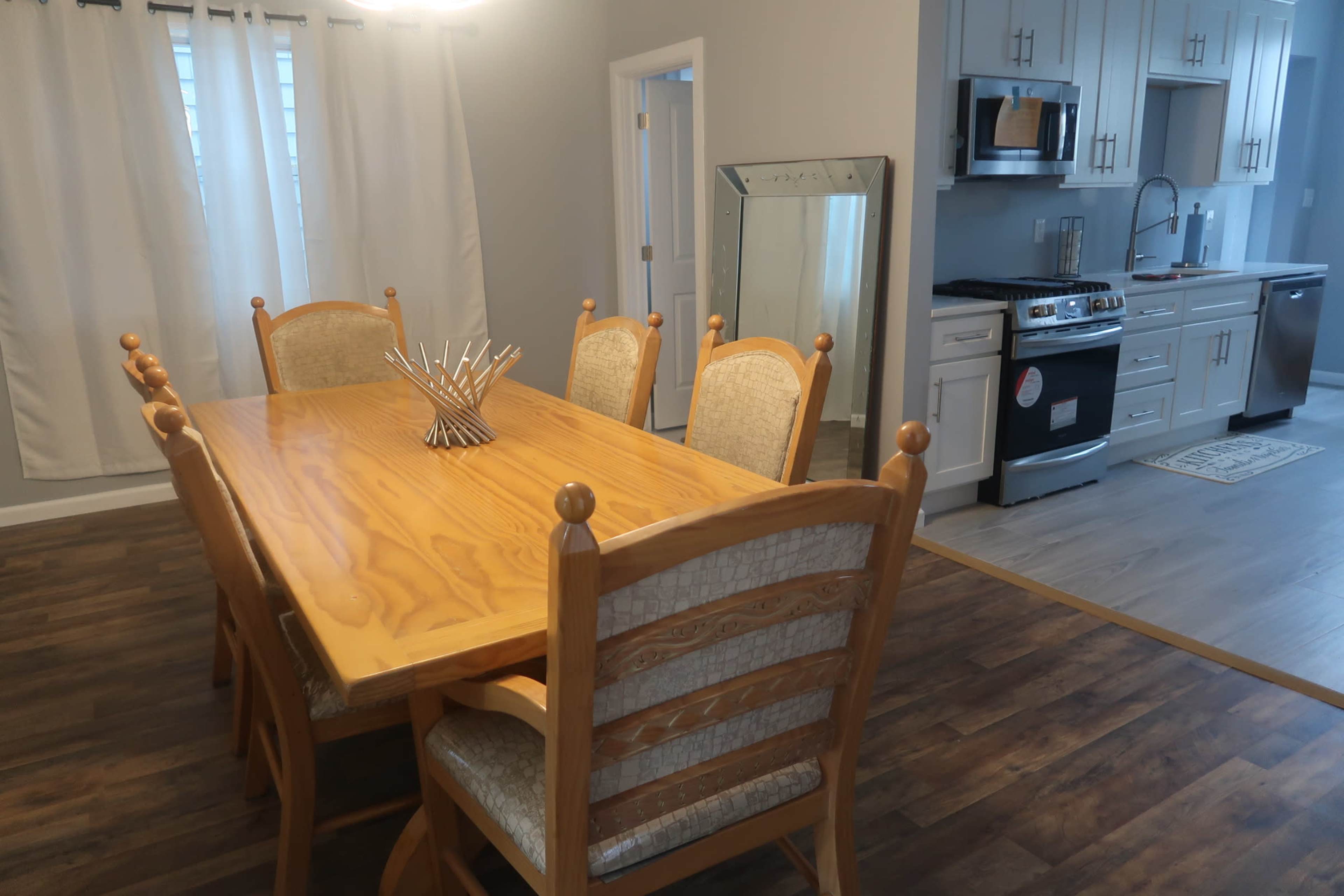 A wooden dining table with eight chairs is positioned beside a kitchen featuring stainless steel appliances and a mirror against the wall.