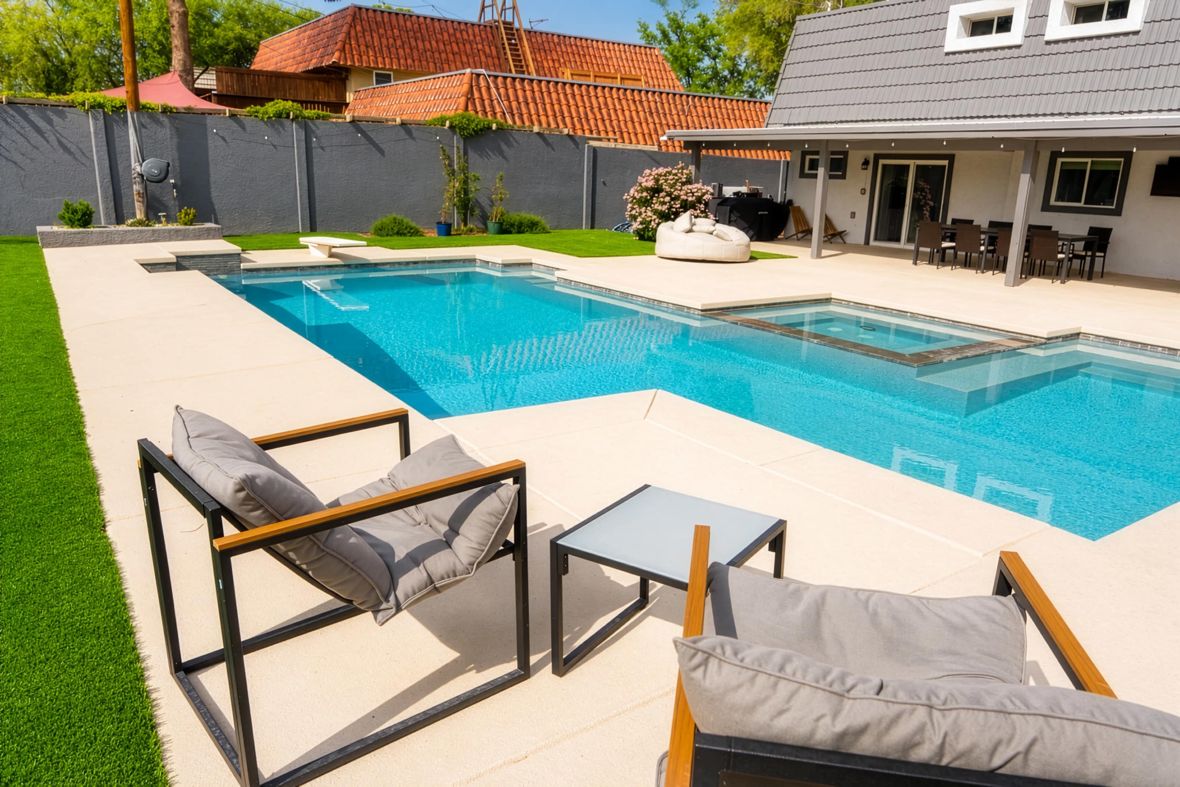 The image shows a modern backyard featuring a blue swimming pool bordered by a patio and stylish lounge chairs.