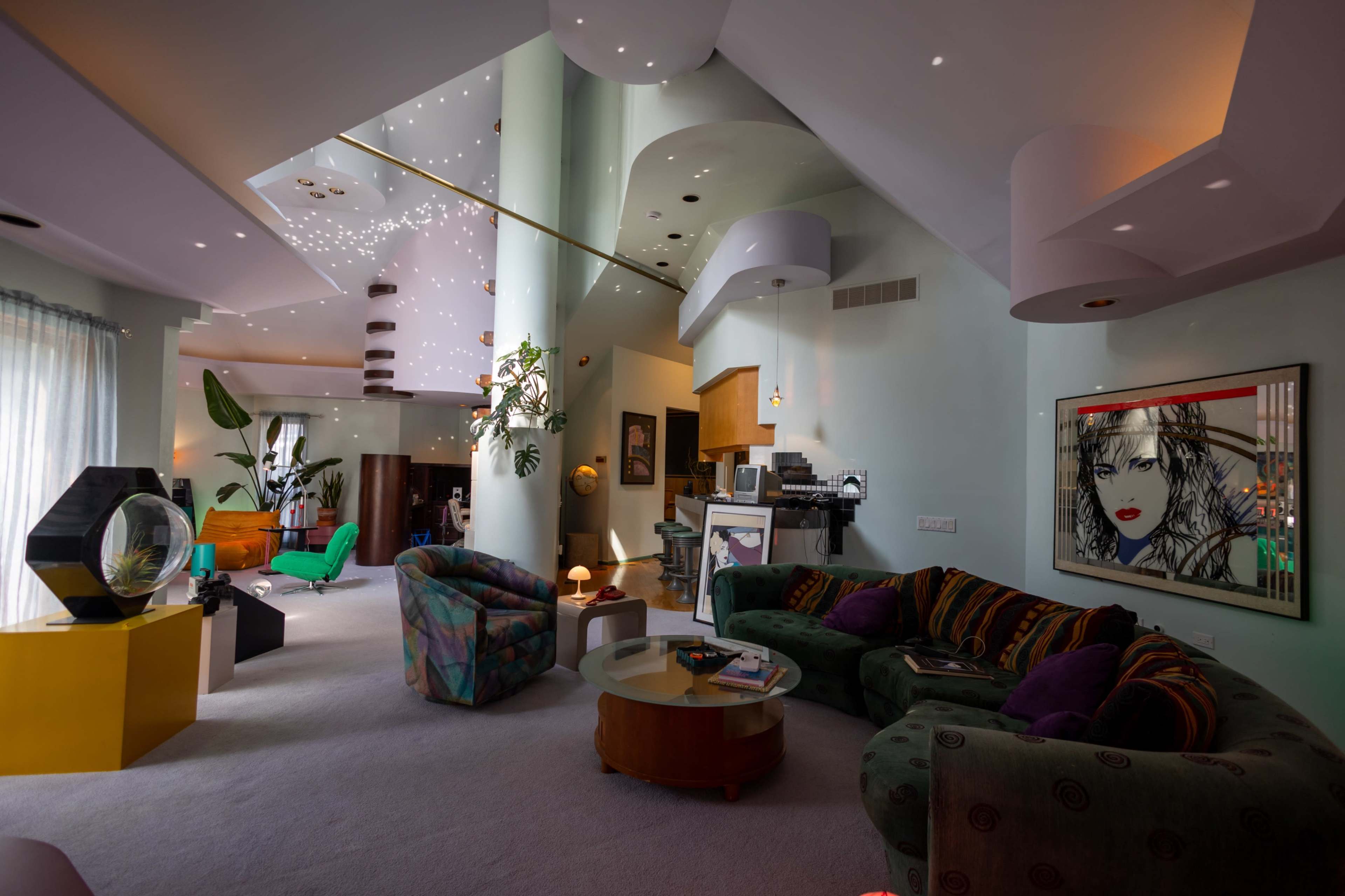 The image shows a spacious living room with distinct architectural features, colorful furniture, and vibrant artwork on the walls.