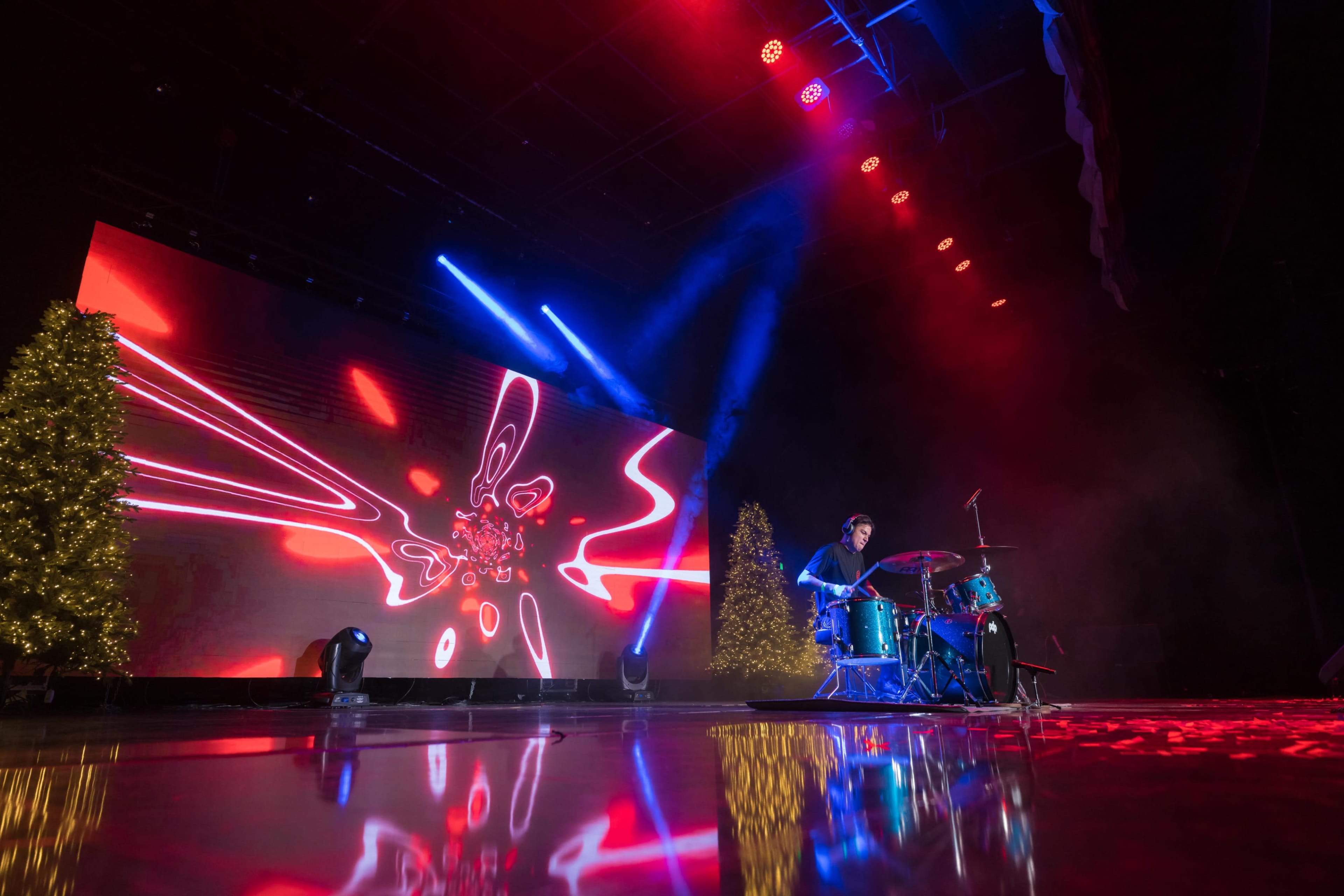 A drummer performs on stage in front of a large illuminated screen with vibrant patterns and two decorated trees.