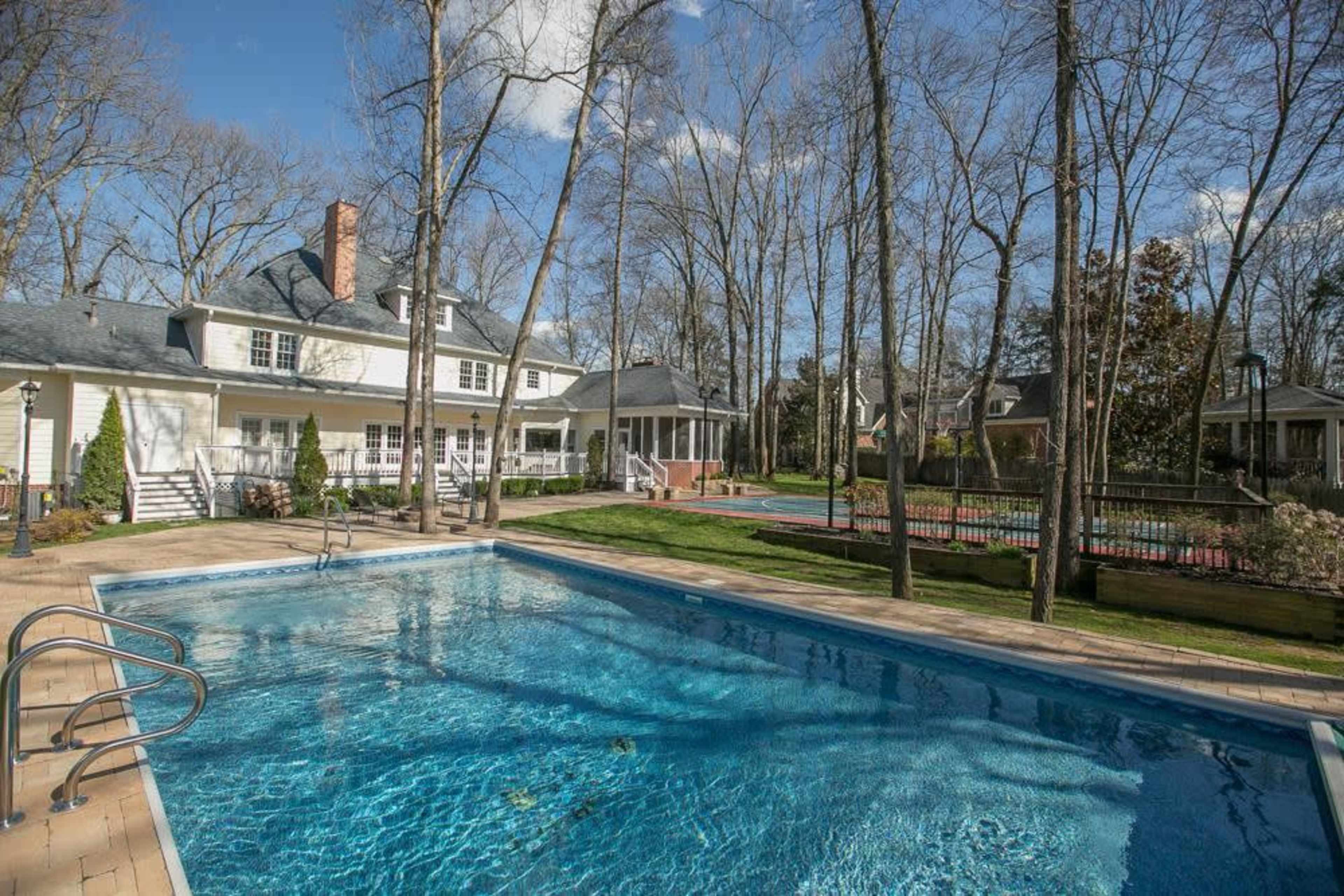 A backyard with a swimming pool surrounded by trees, a house in the background, and a tennis court to the side.