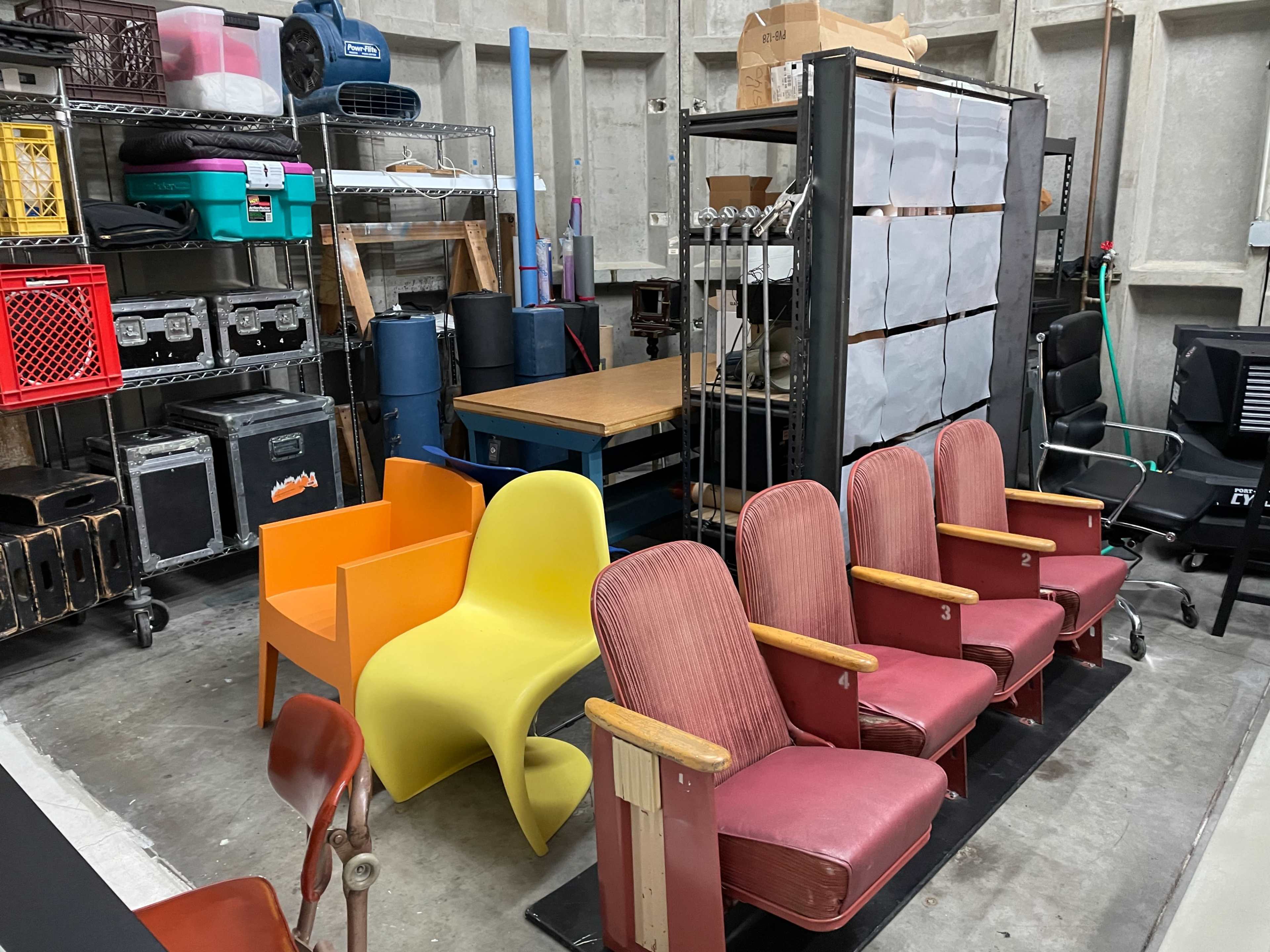 A storage area filled with various chairs, including bright yellow and orange modern chairs alongside red and maroon vintage seating, surrounded by shelves full of boxes and equipment.