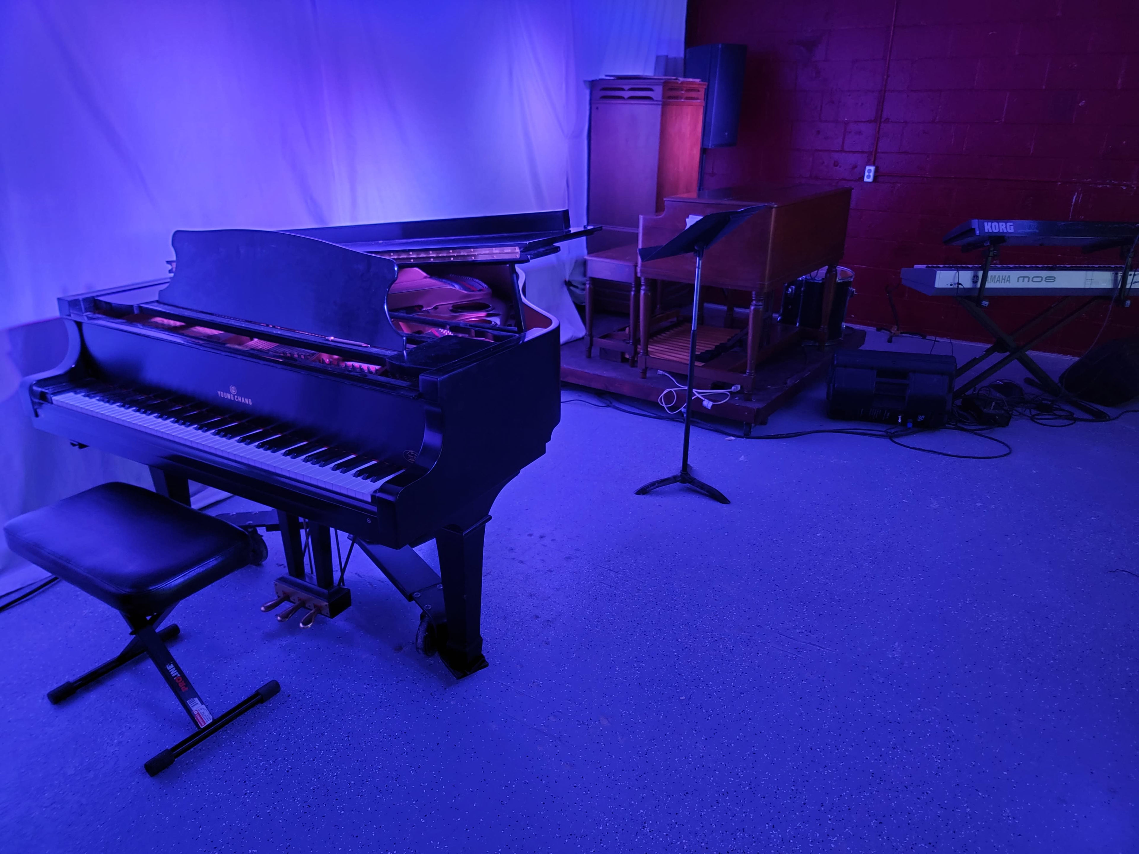 A black grand piano is positioned beside an organ and a keyboard in a room illuminated with blue light.
