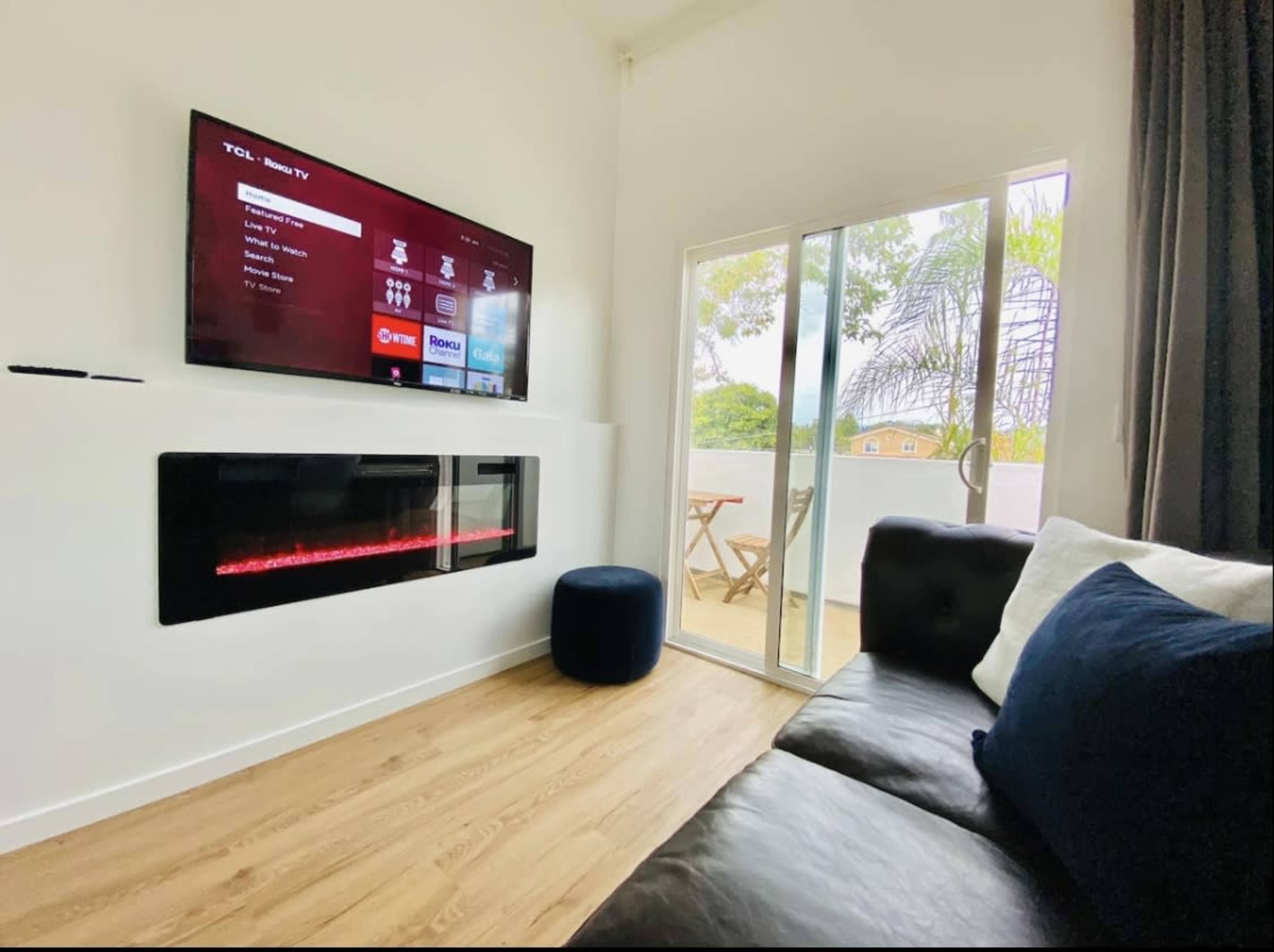 A modern living room with a wall-mounted TV and an electric fireplace beneath it, adjacent to a sliding glass door leading to a small outdoor balcony.