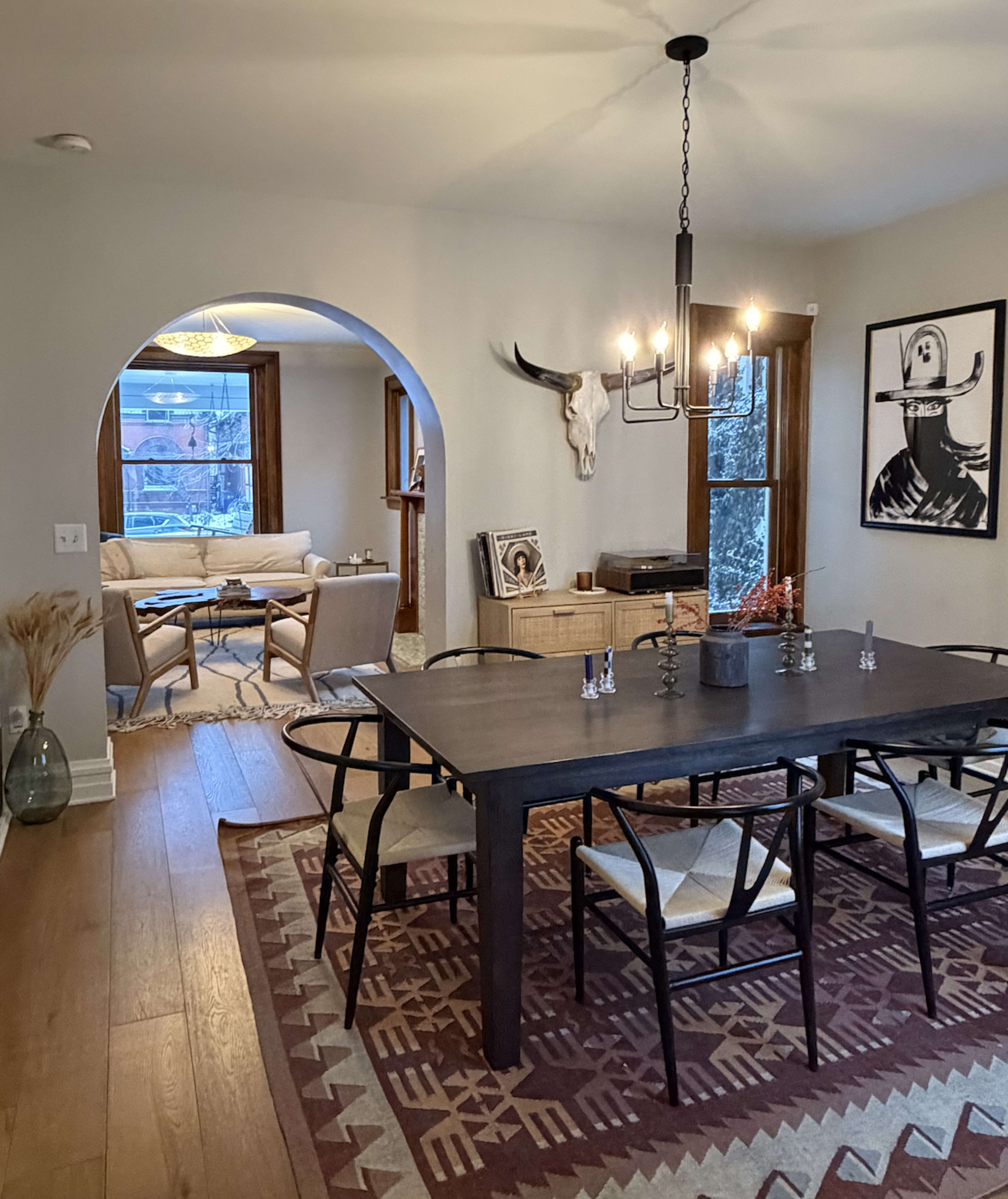 A dining room features a wooden table surrounded by chairs, with an archway leading to a cozy living area illuminated by natural light.