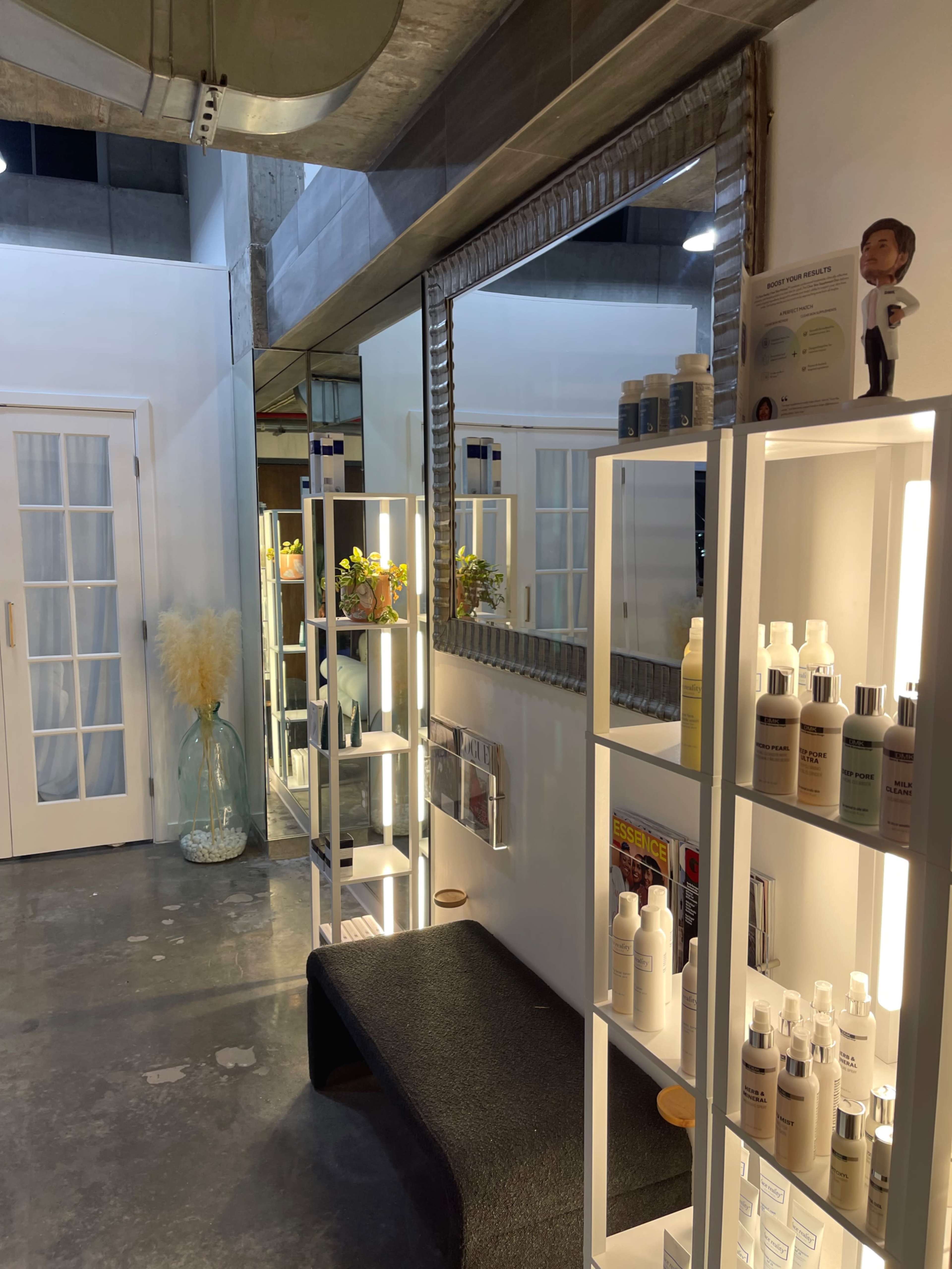 The image shows a modern salon interior with white walls, a large mirror, and shelves displaying various beauty products.