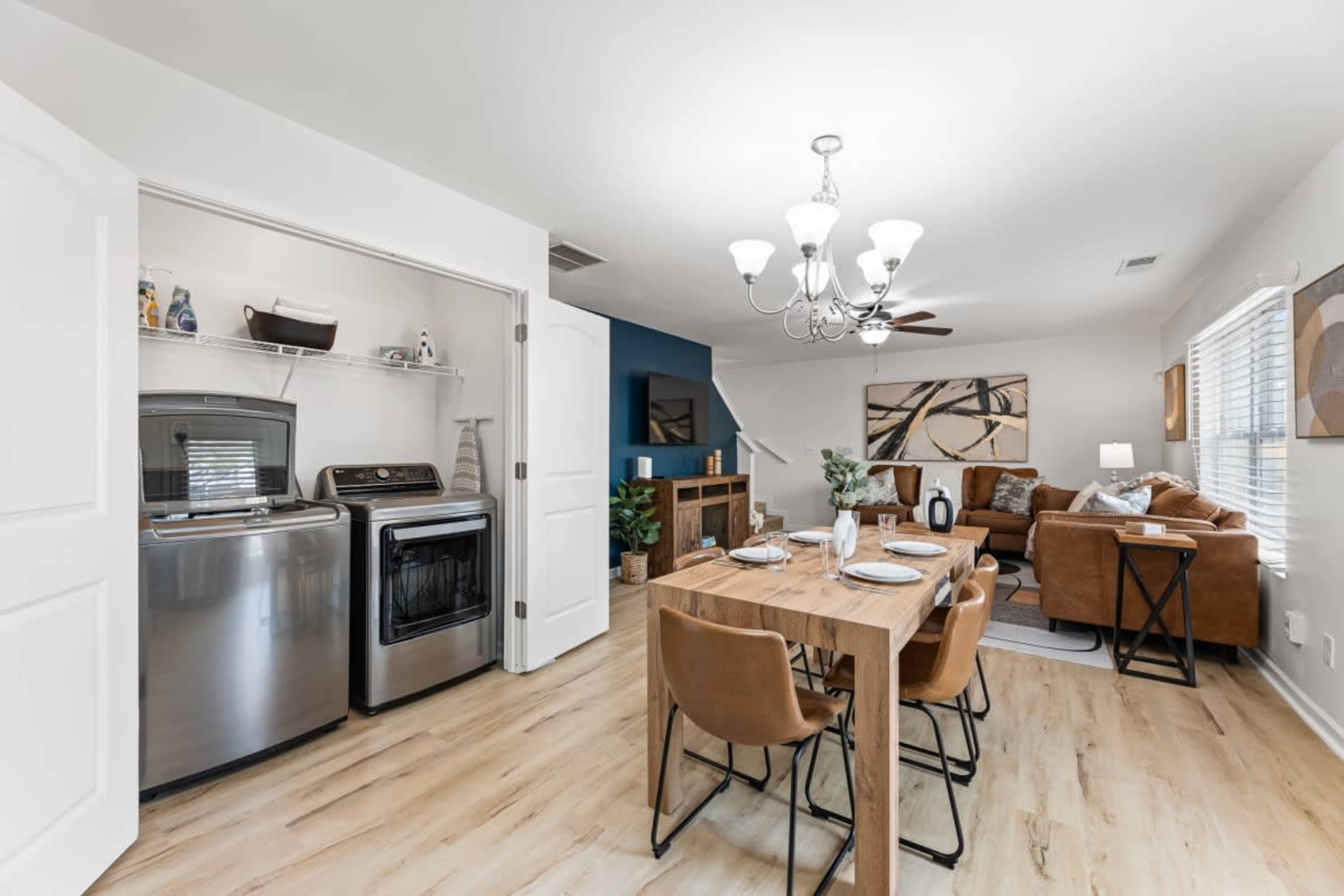 A laundry area is visible next to a dining space that features a wooden table, chairs, and a chandelier, with a living area in the background.