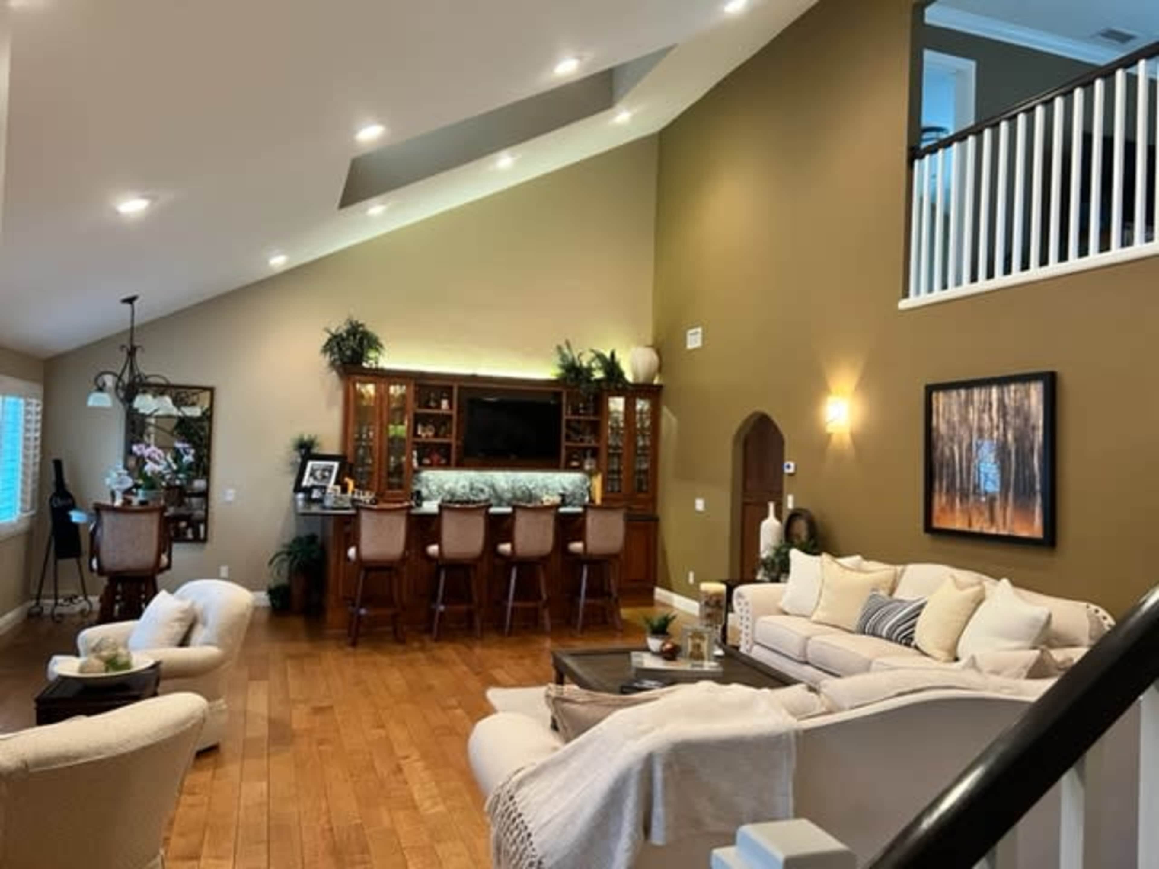 A spacious living area featuring a bar with seating, a light-colored sofa, and large windows, all set against a backdrop of muted wall colors and wooden flooring.