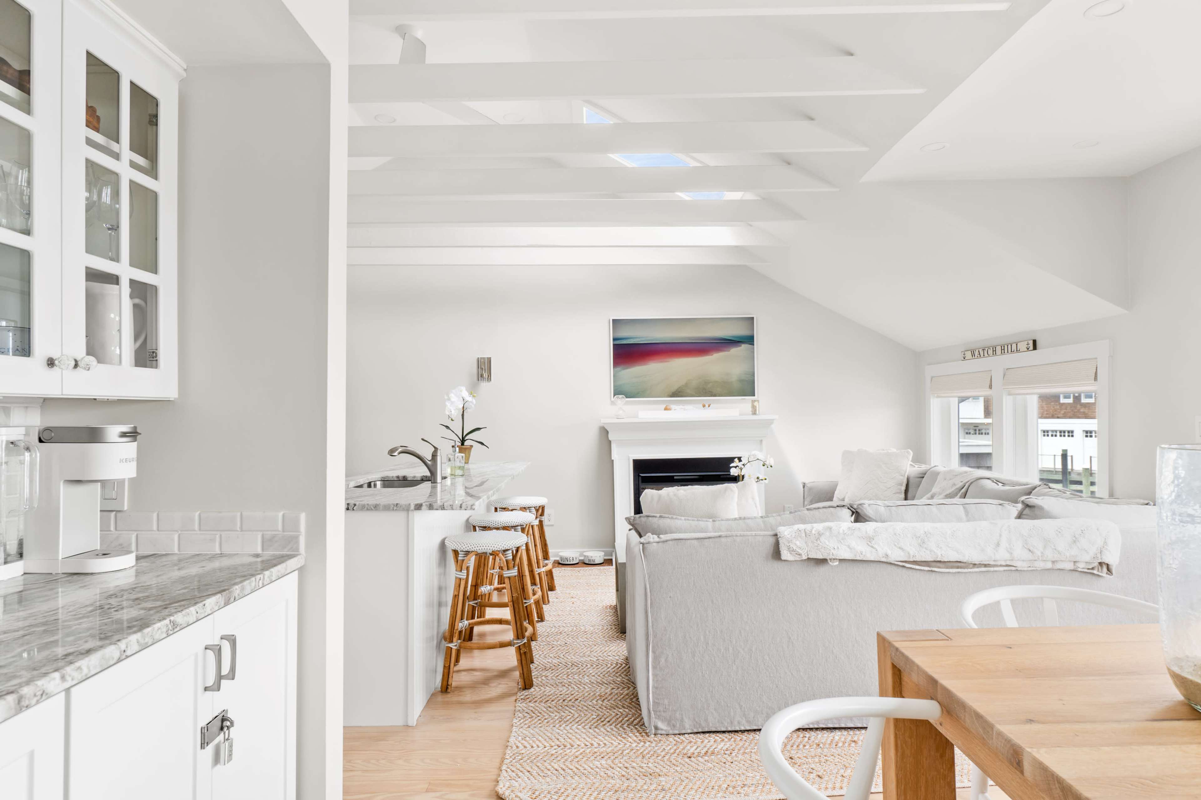 A bright, open living space features a gray sofa, a kitchen with bar stools, and large windows letting in natural light.