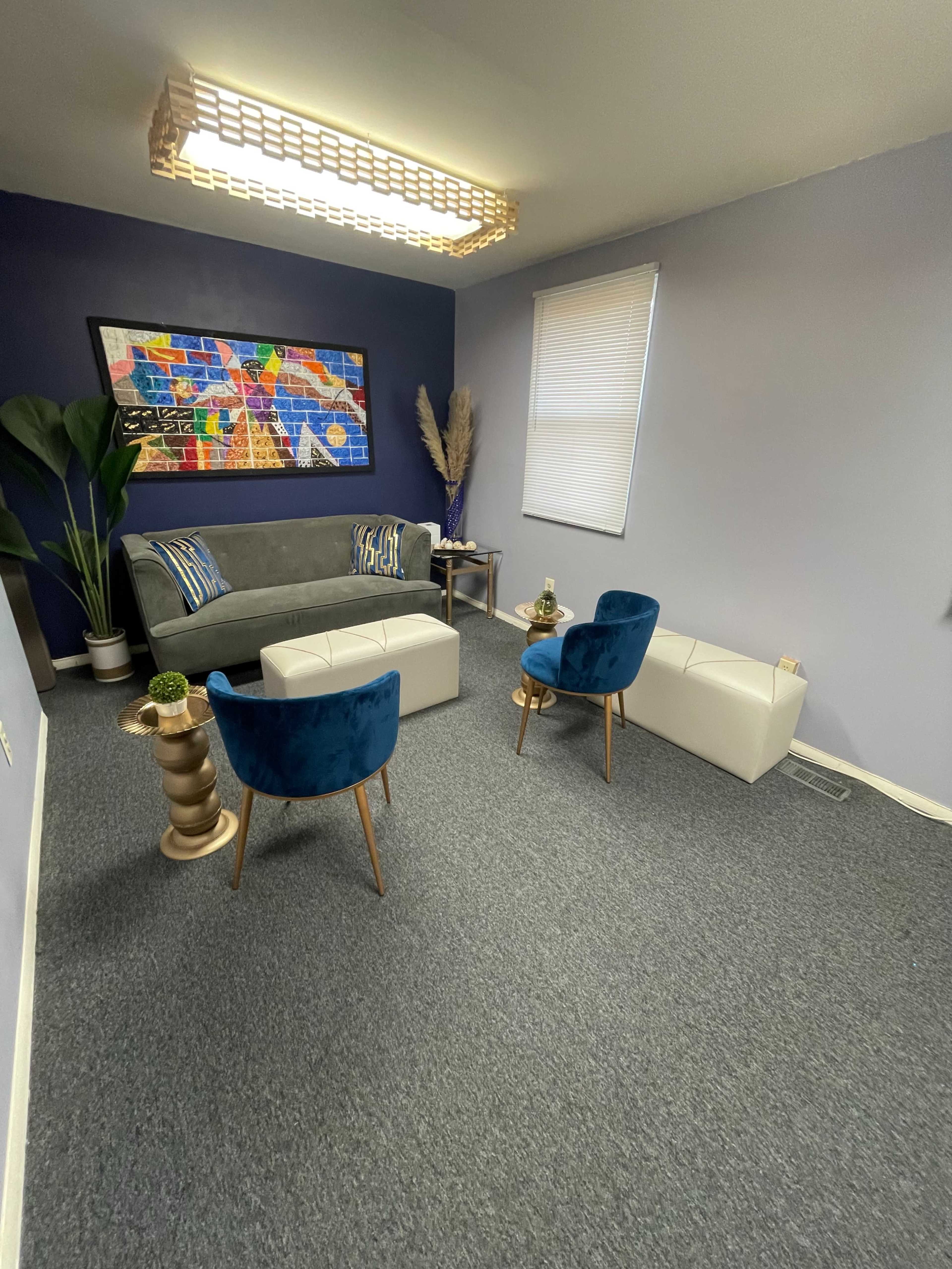 A small room features a gray sofa, two blue velvet chairs, a white coffee table, and colorful wall art illuminated by a modern light fixture.