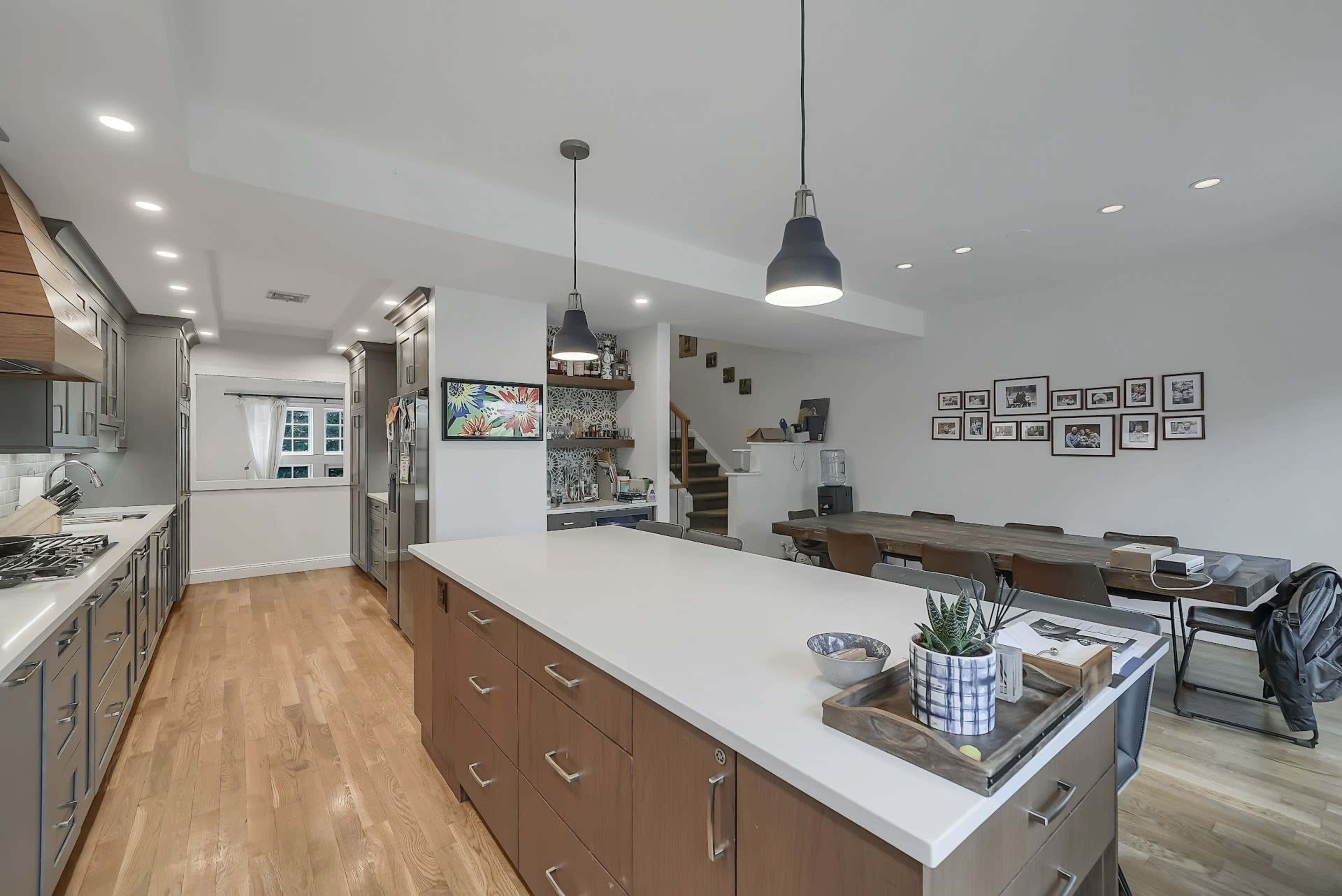 A modern kitchen with an island, stainless steel appliances, and a dining area featuring a long wooden table and a gallery wall of framed pictures.