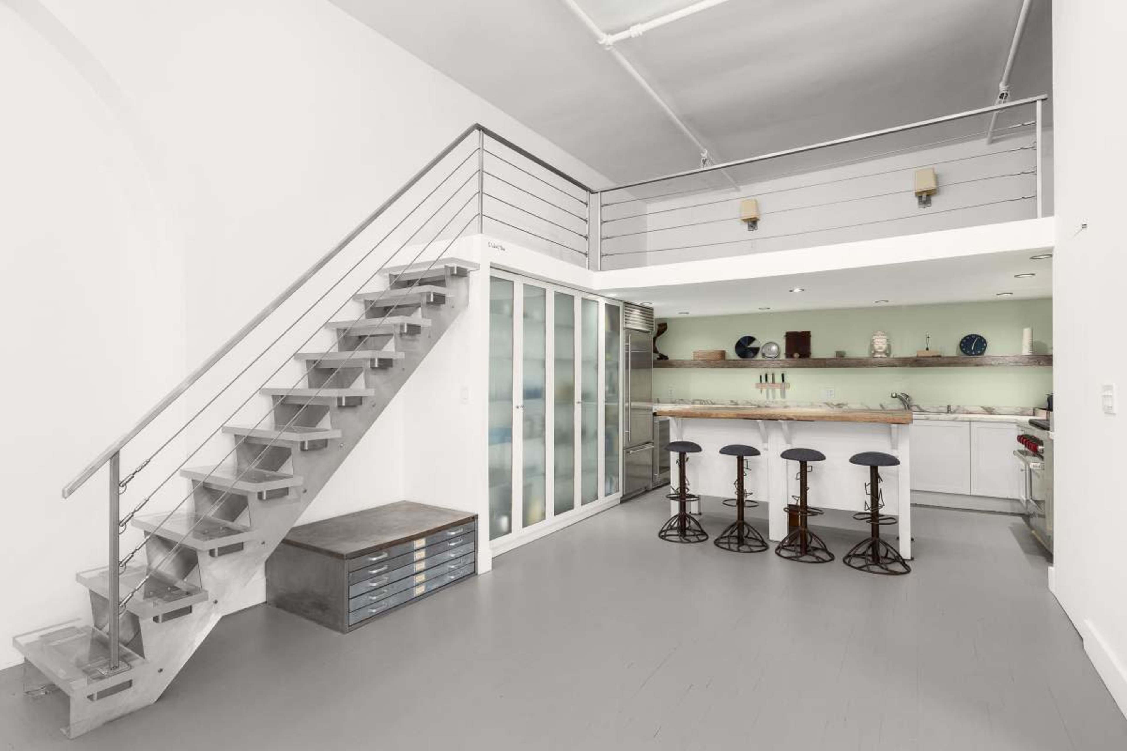The image shows a modern interior space featuring a staircase leading to an upper level, a kitchen area with bar stools, and a minimalist design.