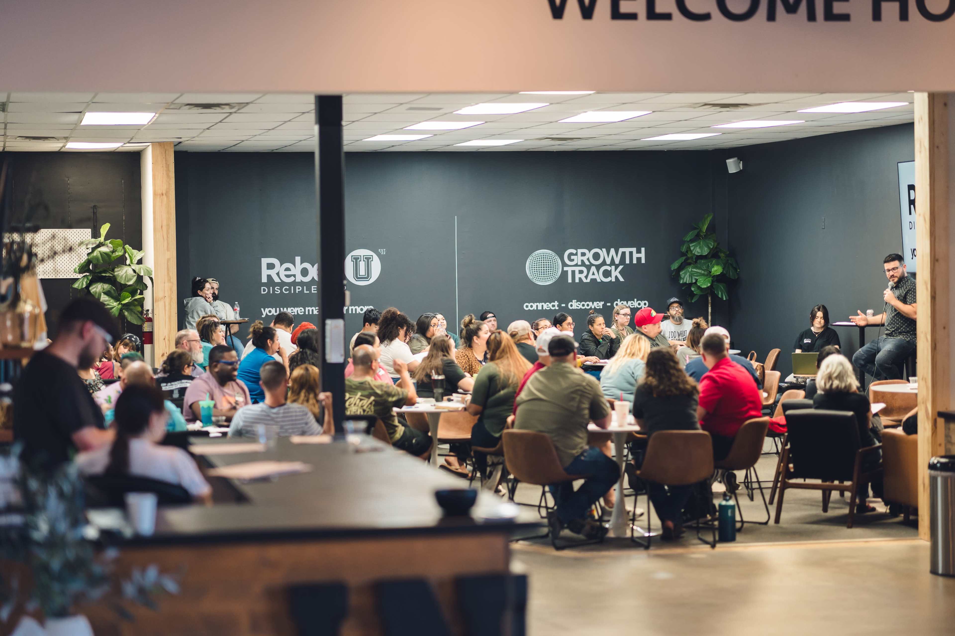 A large group of people sits around tables in a meeting room, engaging in discussion while a speaker addresses them from the front.