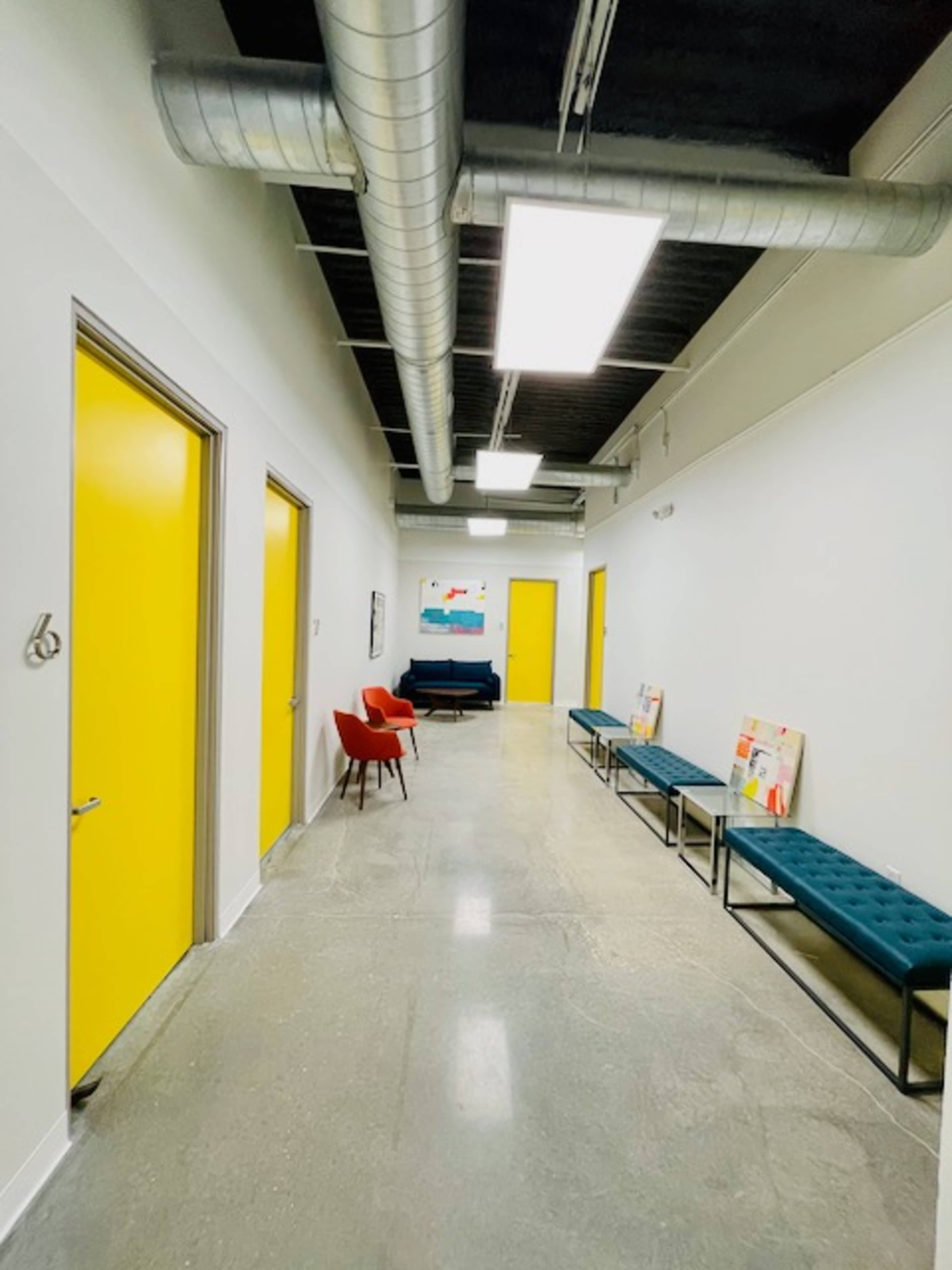 A hallway features yellow doors, benches with teal upholstery, and a red chair, illuminated by overhead lights.