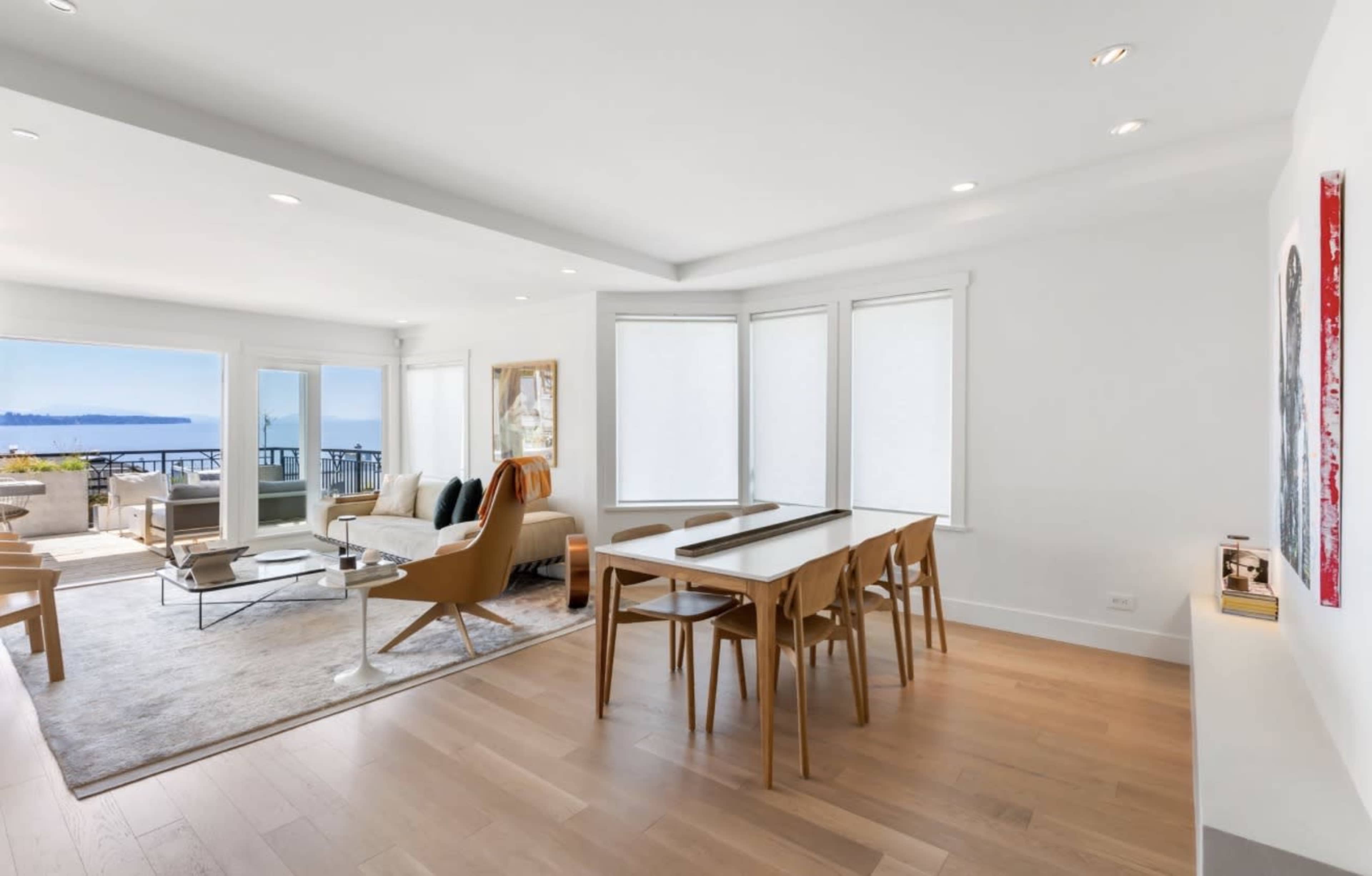 The image shows a modern living space with a dining table, comfortable seating area, and large windows offering views of the outdoors.