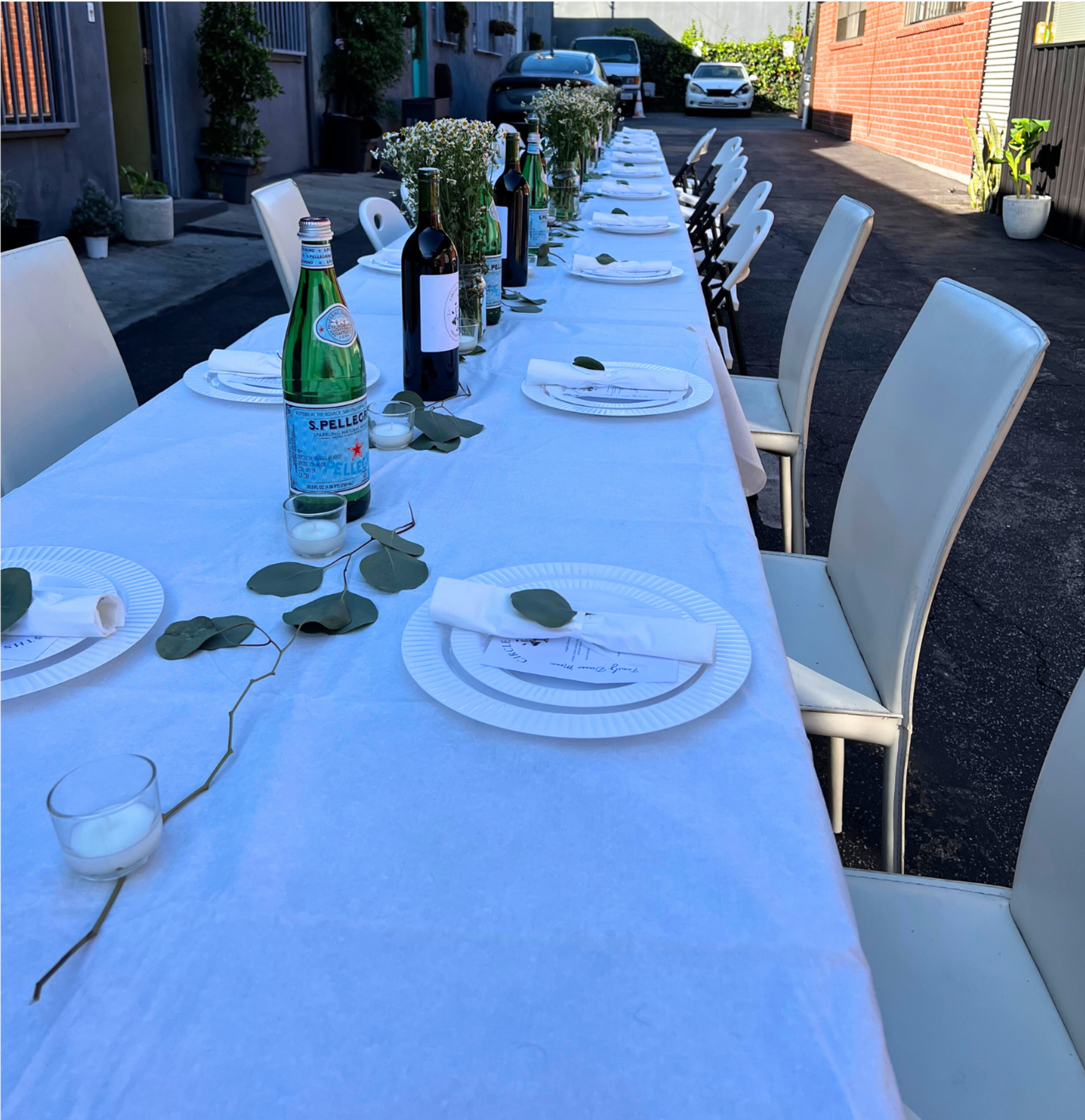 A long table is set for a meal, adorned with white tablecloths, plates, wine bottles, and decorative greenery, situated in a narrow outdoor space.