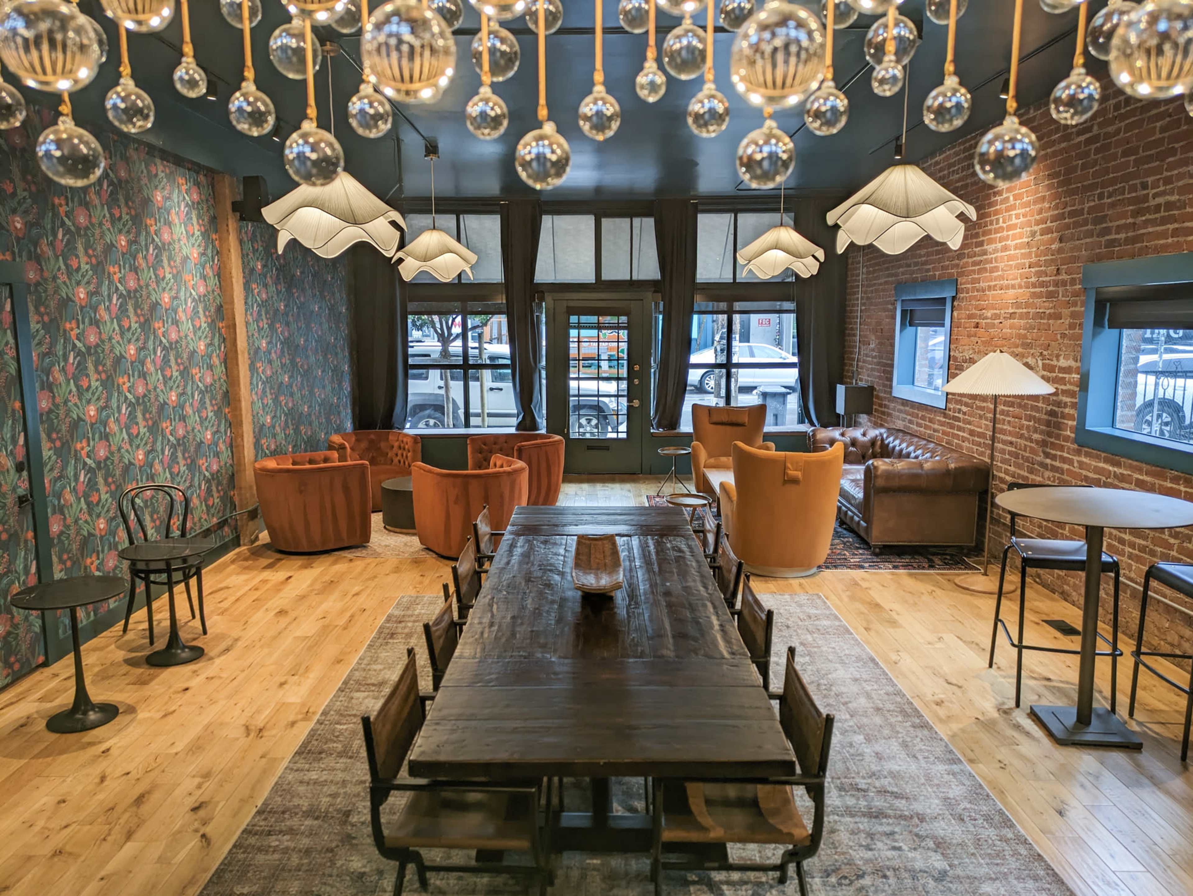 A spacious, modern lounge area featuring a long wooden table, several seating arrangements with armchairs, and unique light fixtures hanging from the ceiling.