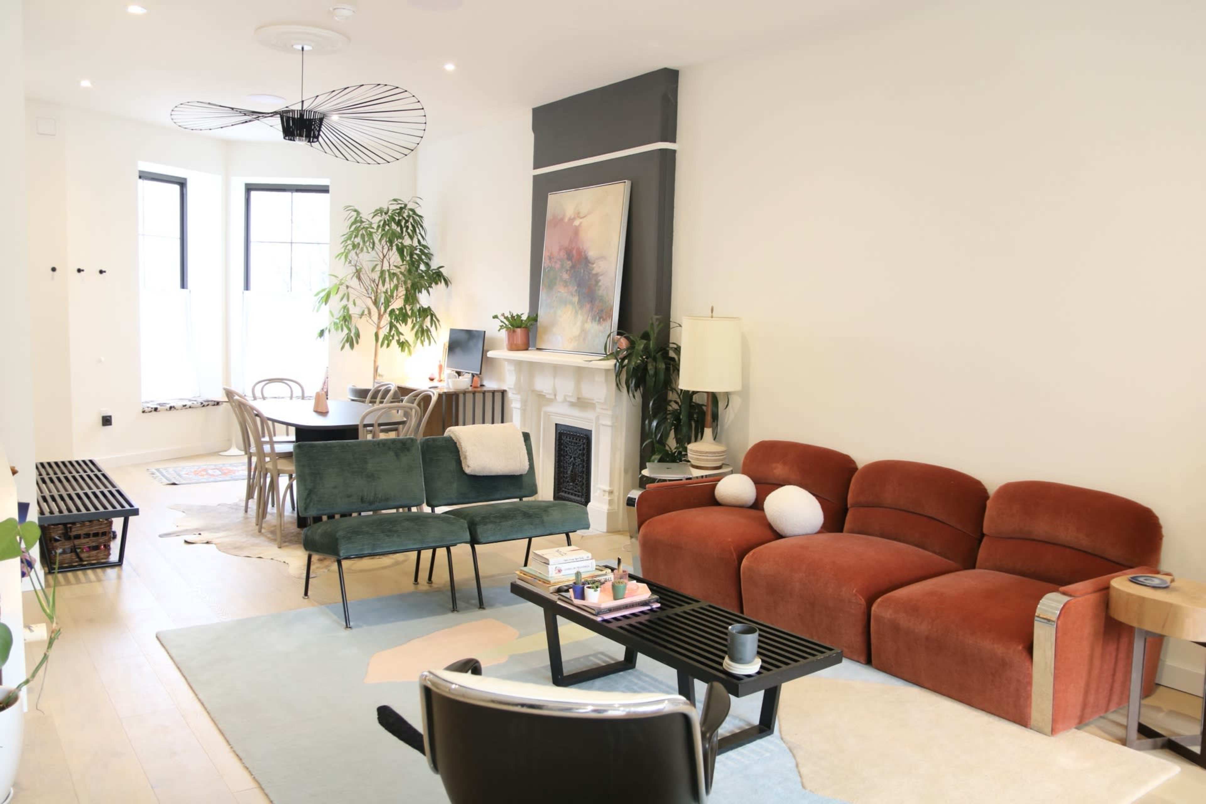 The image shows a modern living room featuring a reddish-brown sectional sofa, a green bench, a round dining table, and decorative plants.