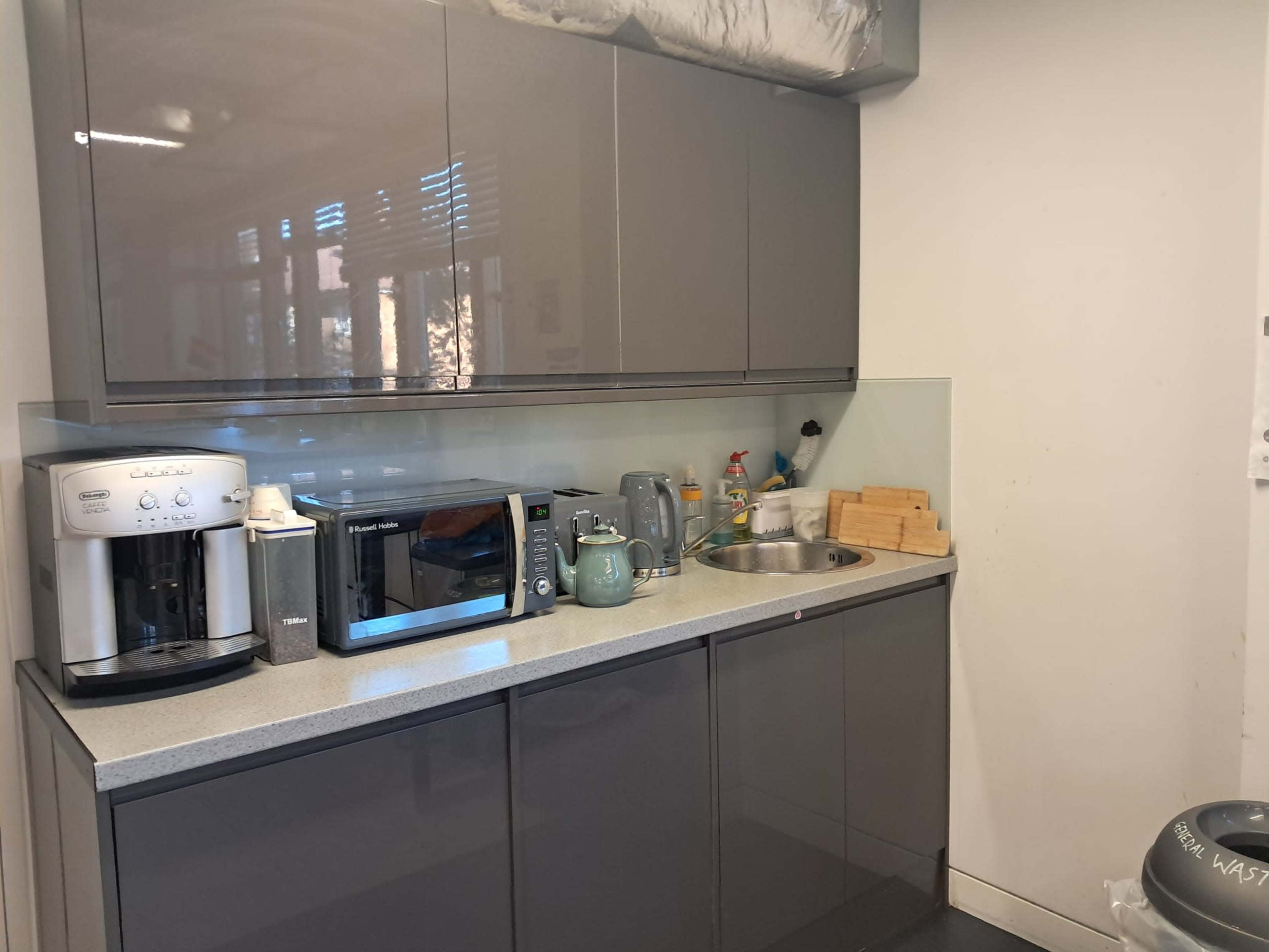 The image shows a modern kitchen corner with gray cabinets, a sink, a microwave, a coffee maker, and various kitchen appliances arranged on the countertop.