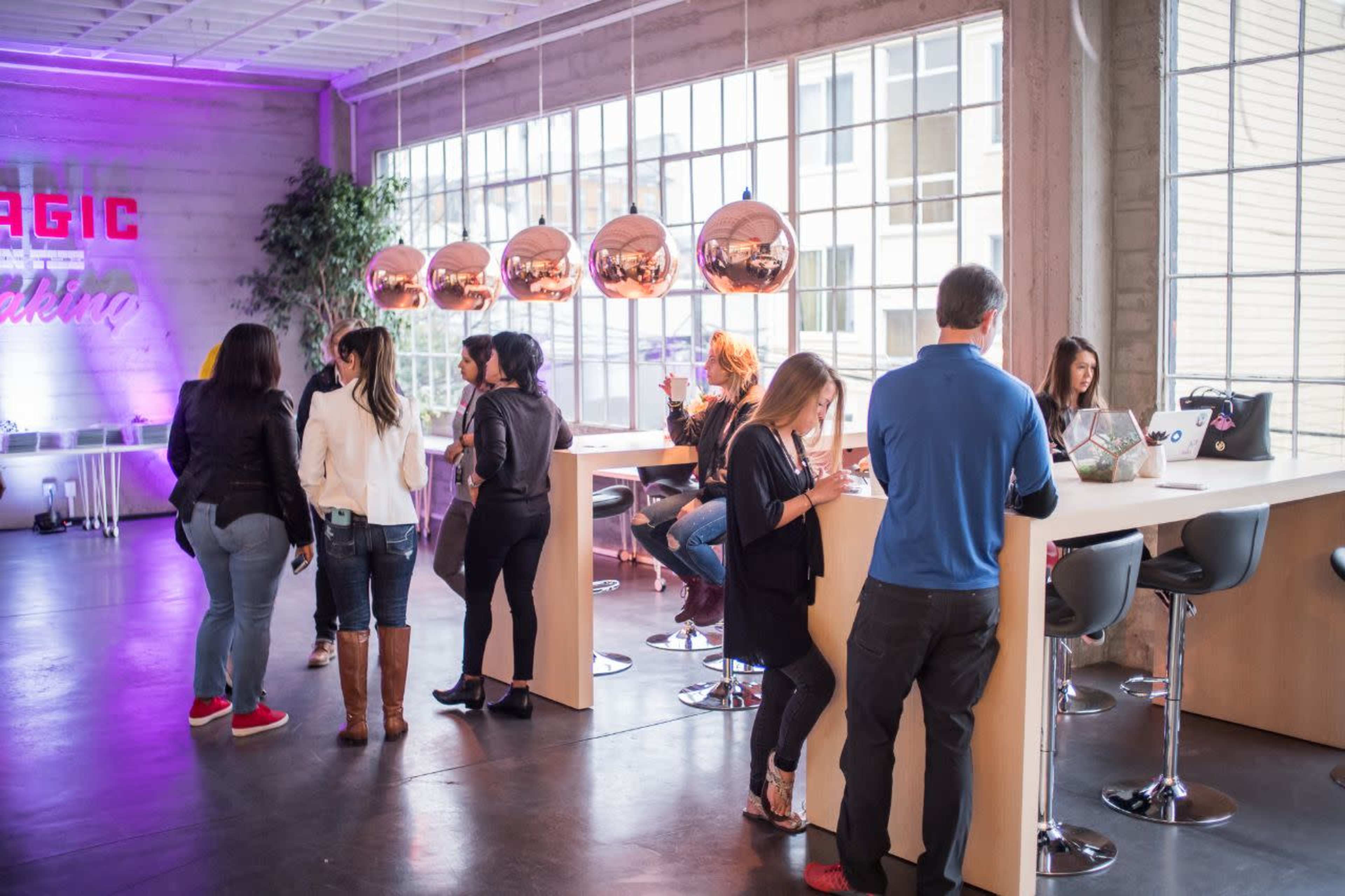 A group of people socializes and works in a modern, well-lit space featuring a long table and decorative hanging lights.