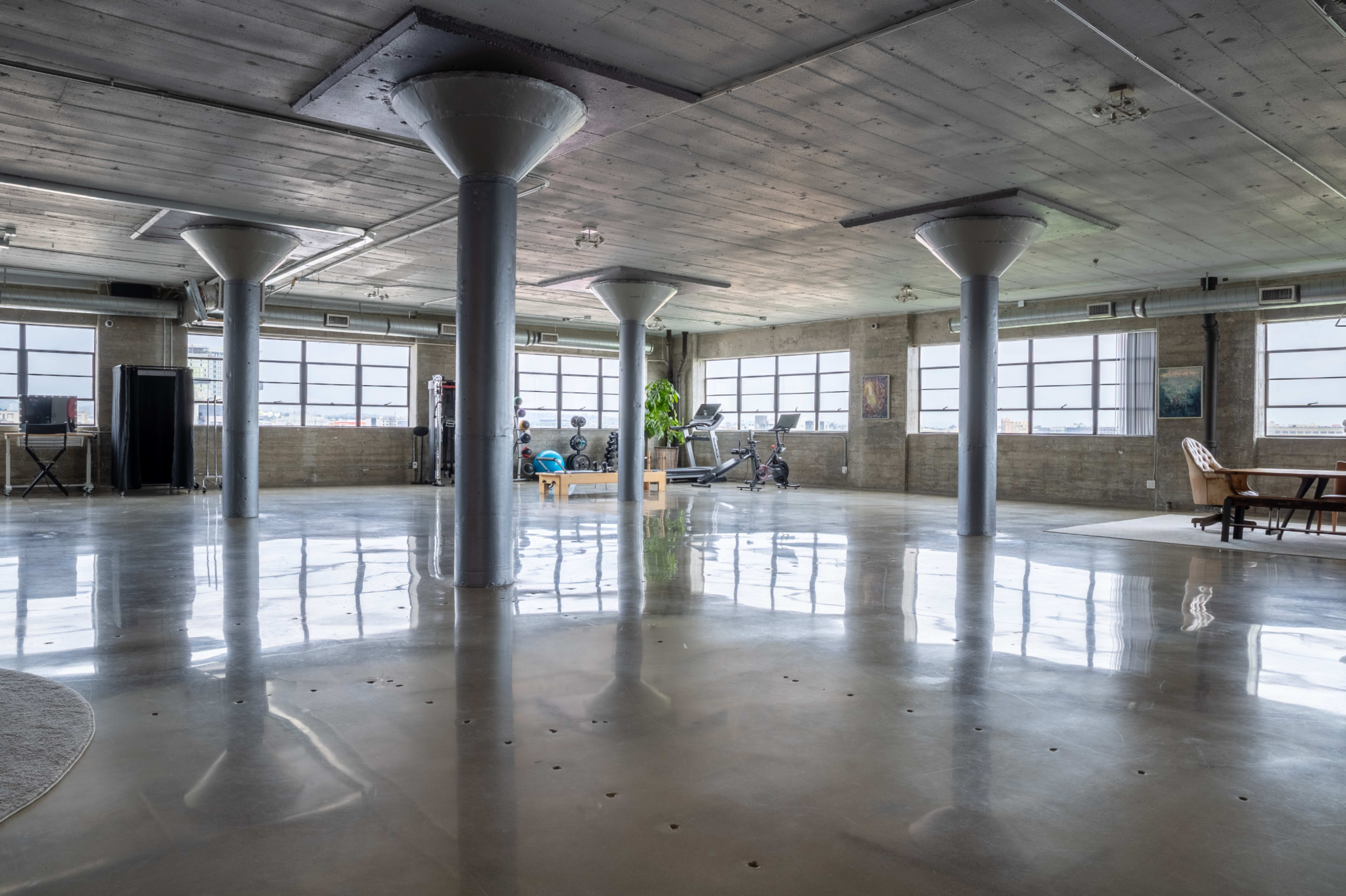 The image depicts a spacious, industrial-style interior with polished concrete floors, large windows, and several columns, featuring workout equipment and a seating area.