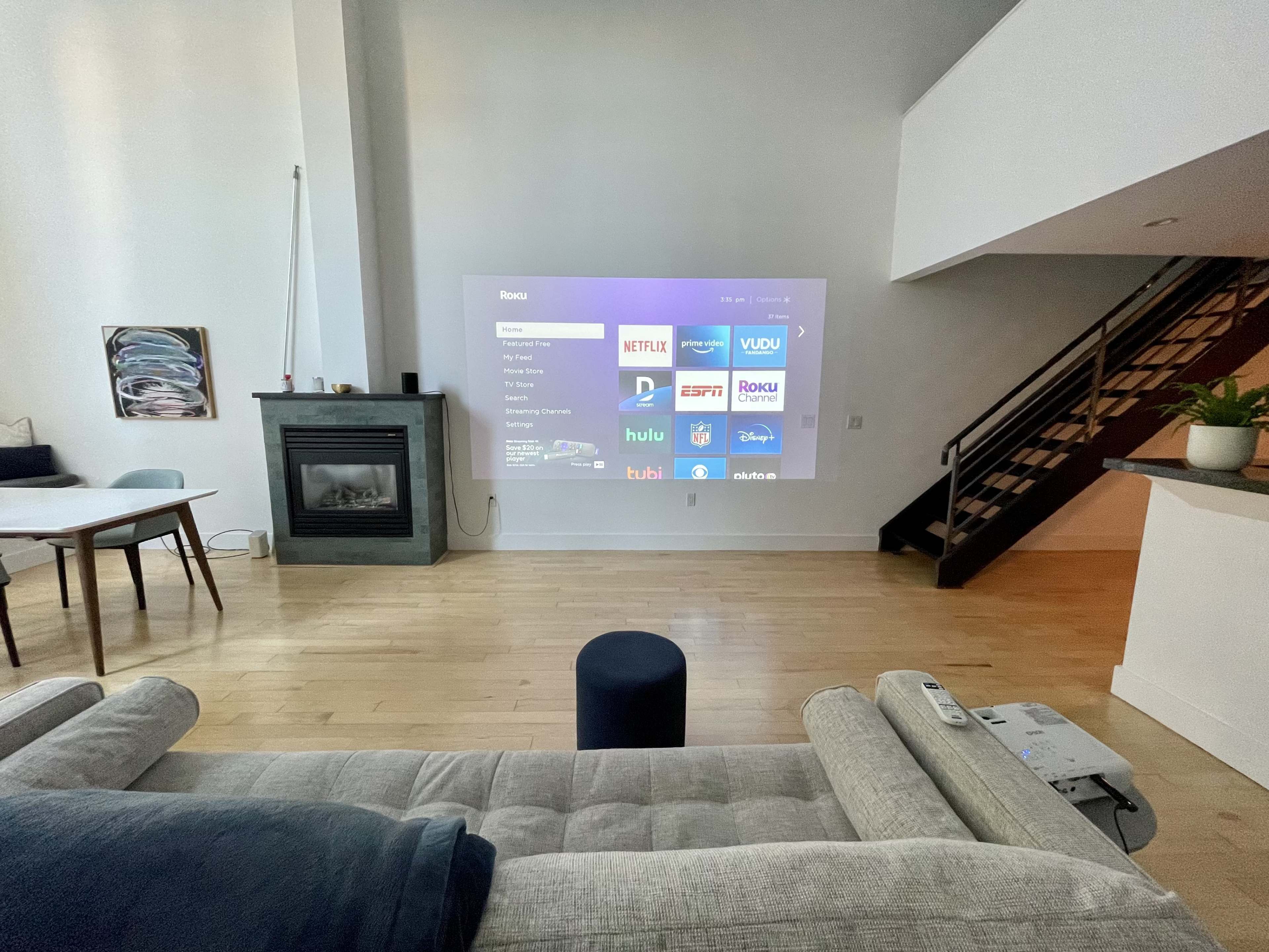 A projector displays a streaming menu on the wall in a modern living room with a fireplace and a staircase.
