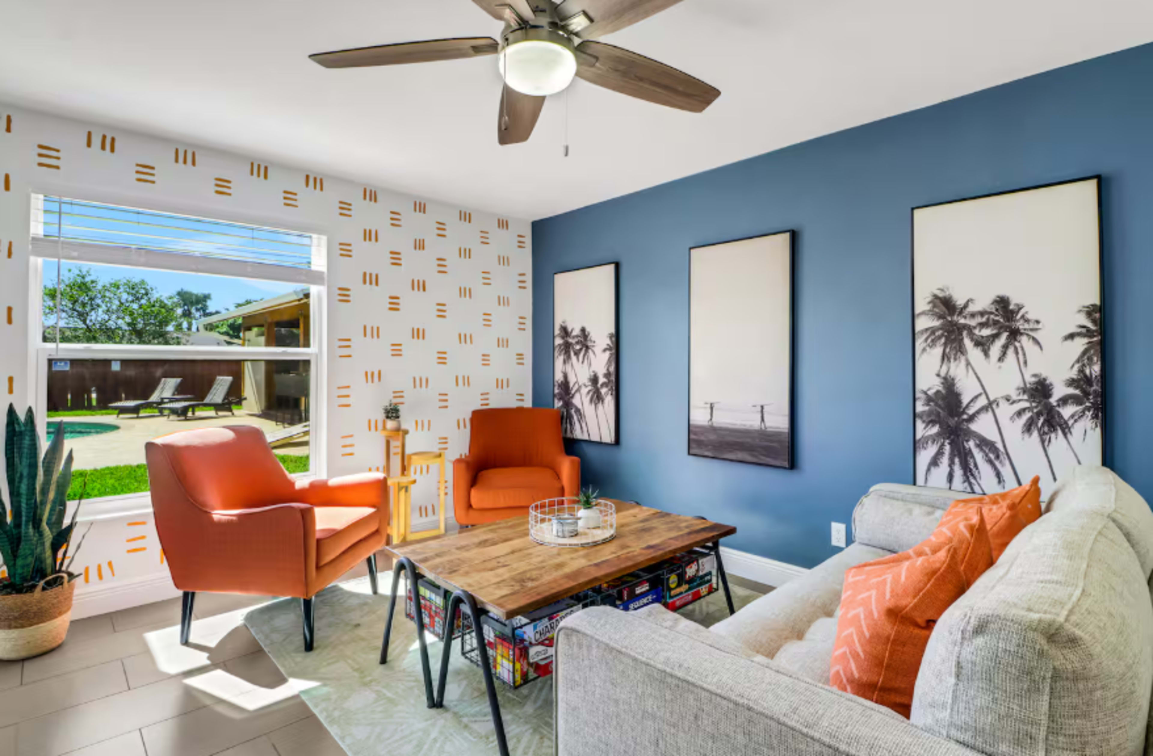 The image shows a spacious living room with a mix of modern decor, featuring two orange chairs, a gray sofa, a wooden coffee table, and wall art depicting palm trees.