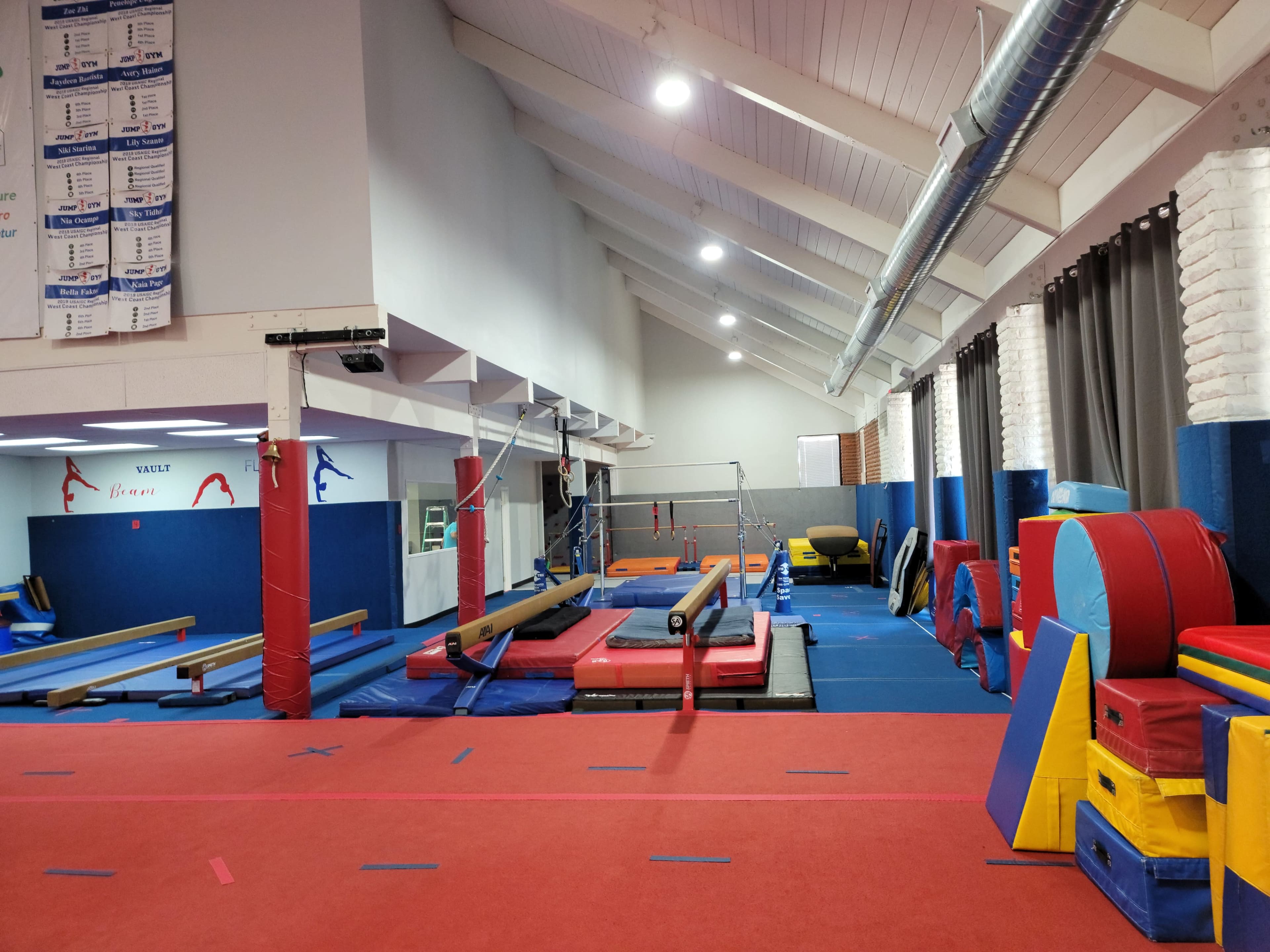 A gymnasium equipped with various gymnastics apparatus, including balance beams, mats, and parallel bars under a high ceiling with bright lighting.