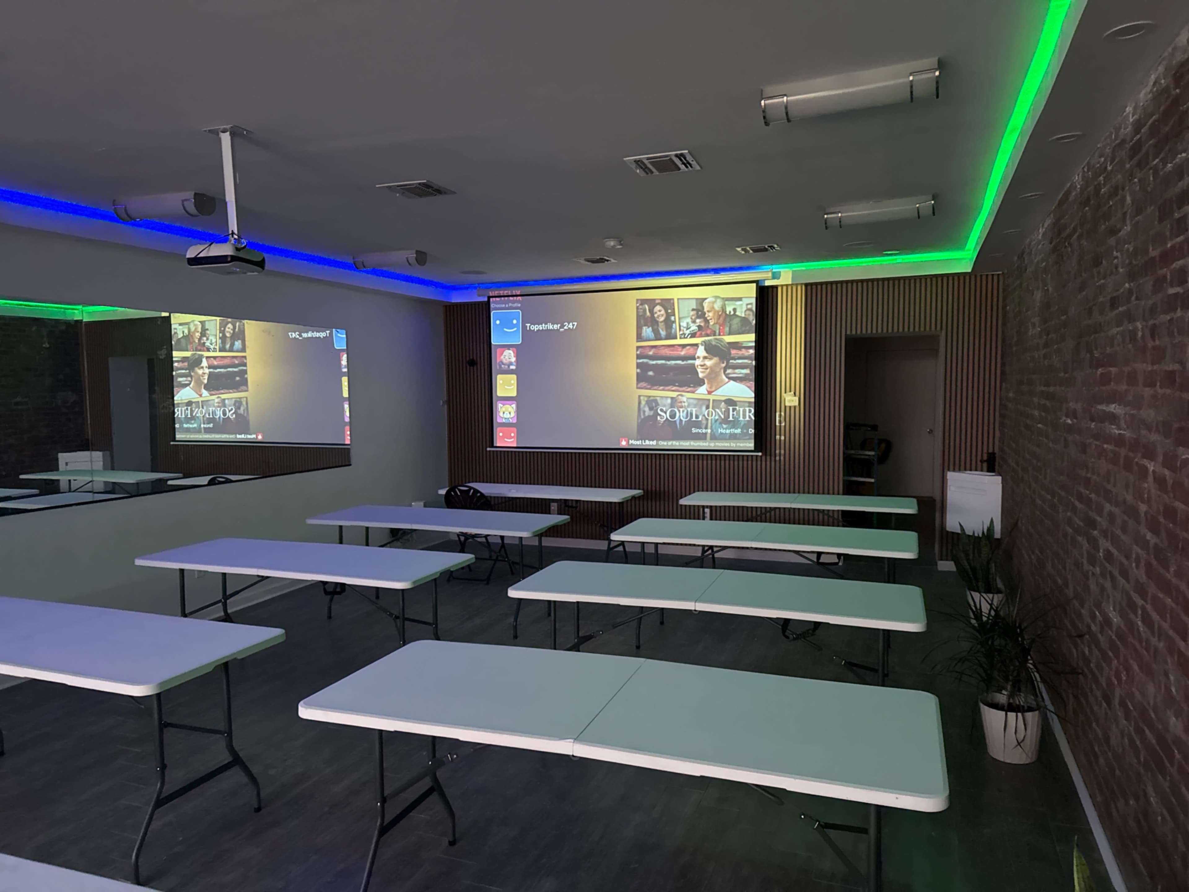 The image shows a modern conference room with multiple white tables arranged in rows, a large projector screen displaying content, and green and blue LED lighting along the walls.