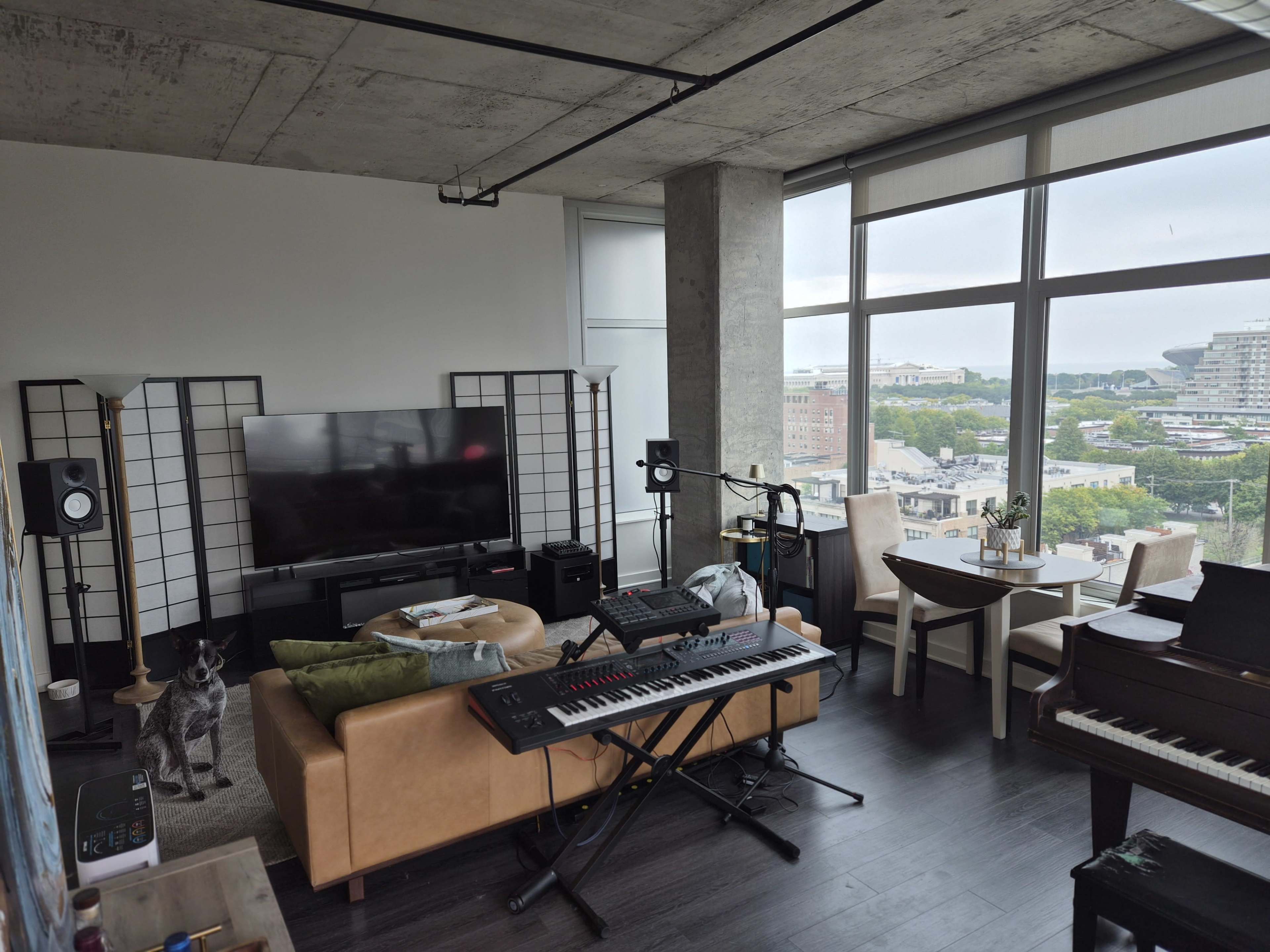 A modern living room features a television, a keyboard, a piano, and a table, with large windows providing a view of the city.
