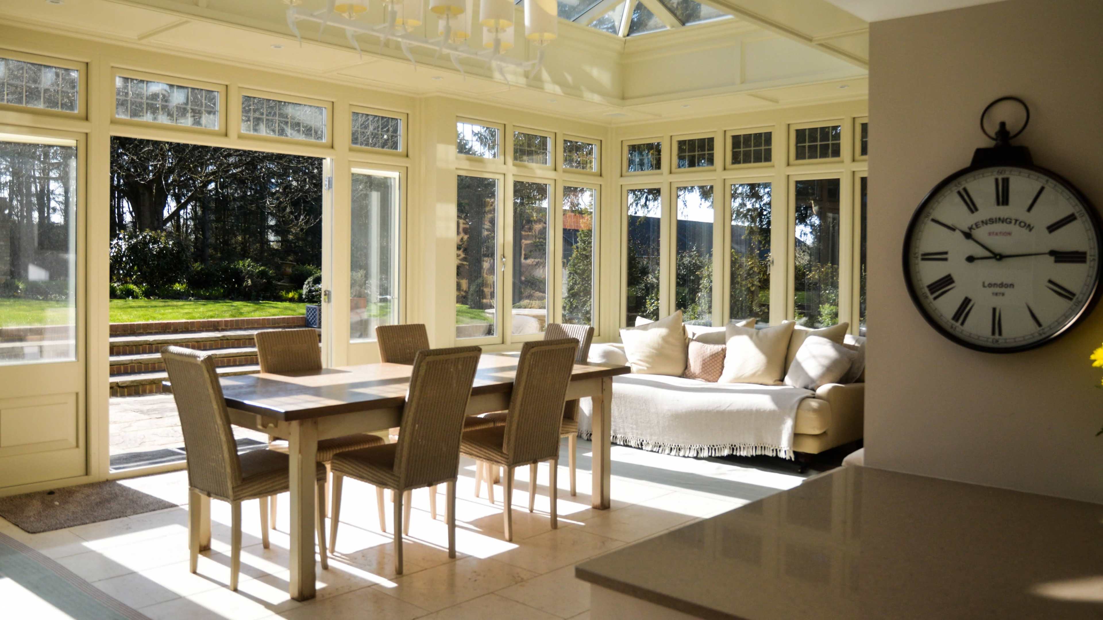A bright sunroom features a wooden dining table with six chairs, a light-colored sofa, and large windows overlooking a garden.