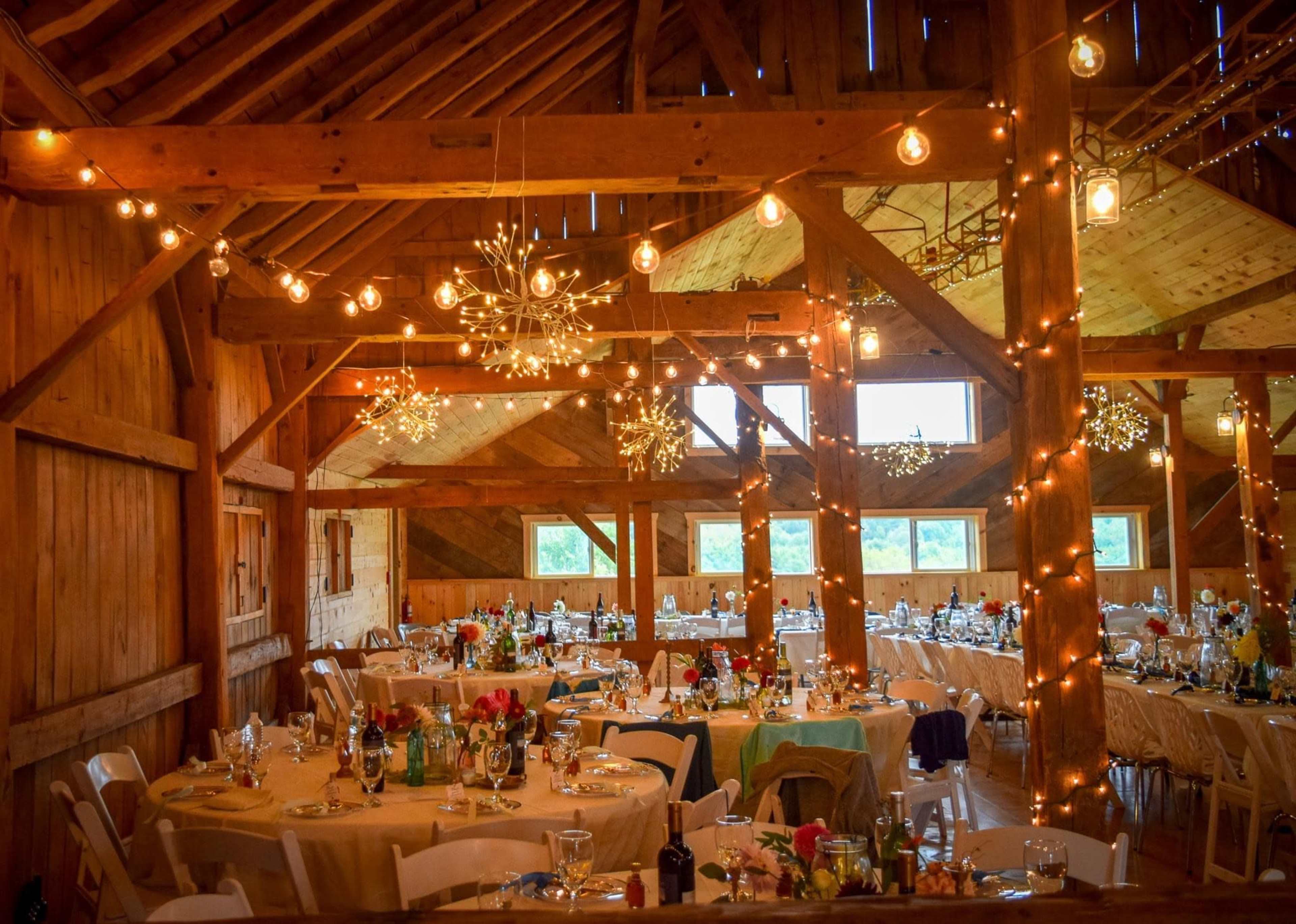 The image shows a beautifully arranged banquet hall with round tables set for a dinner, decorated with flowers and illuminated by string lights and chandeliers.
