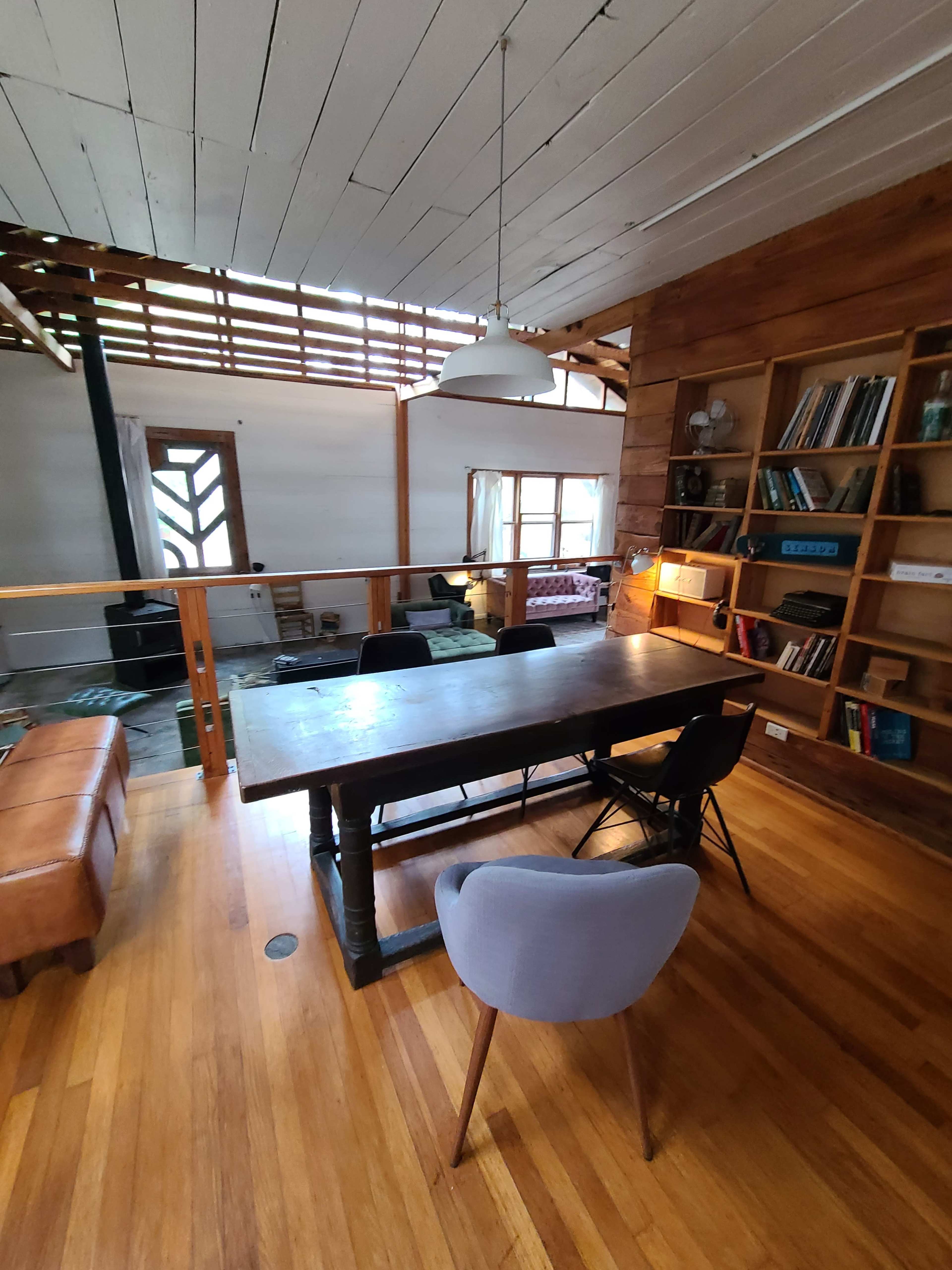 The image shows a spacious, wooden-floored room with a large dining table surrounded by chairs, a couch in the corner, and shelves filled with books along the wall.