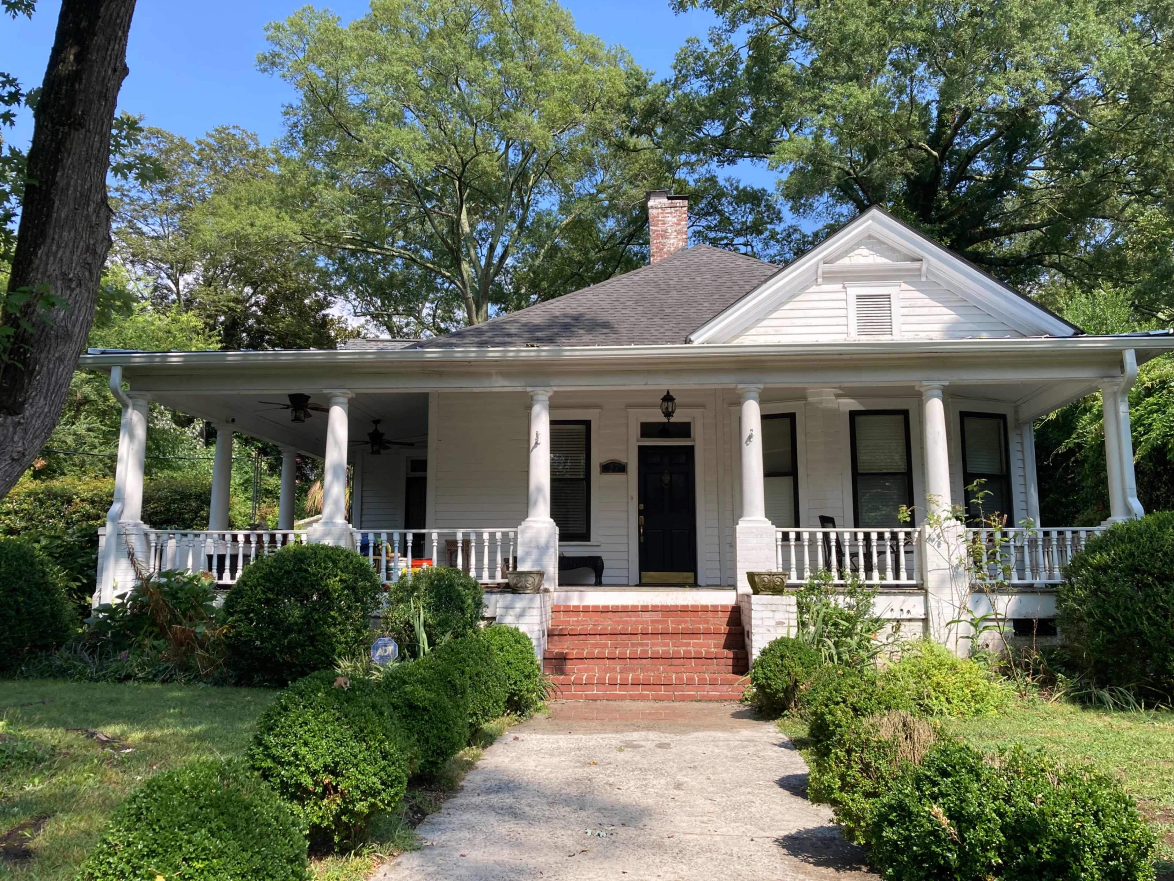 A white, one-story house with a front porch surrounded by neatly trimmed bushes and a brick walkway.