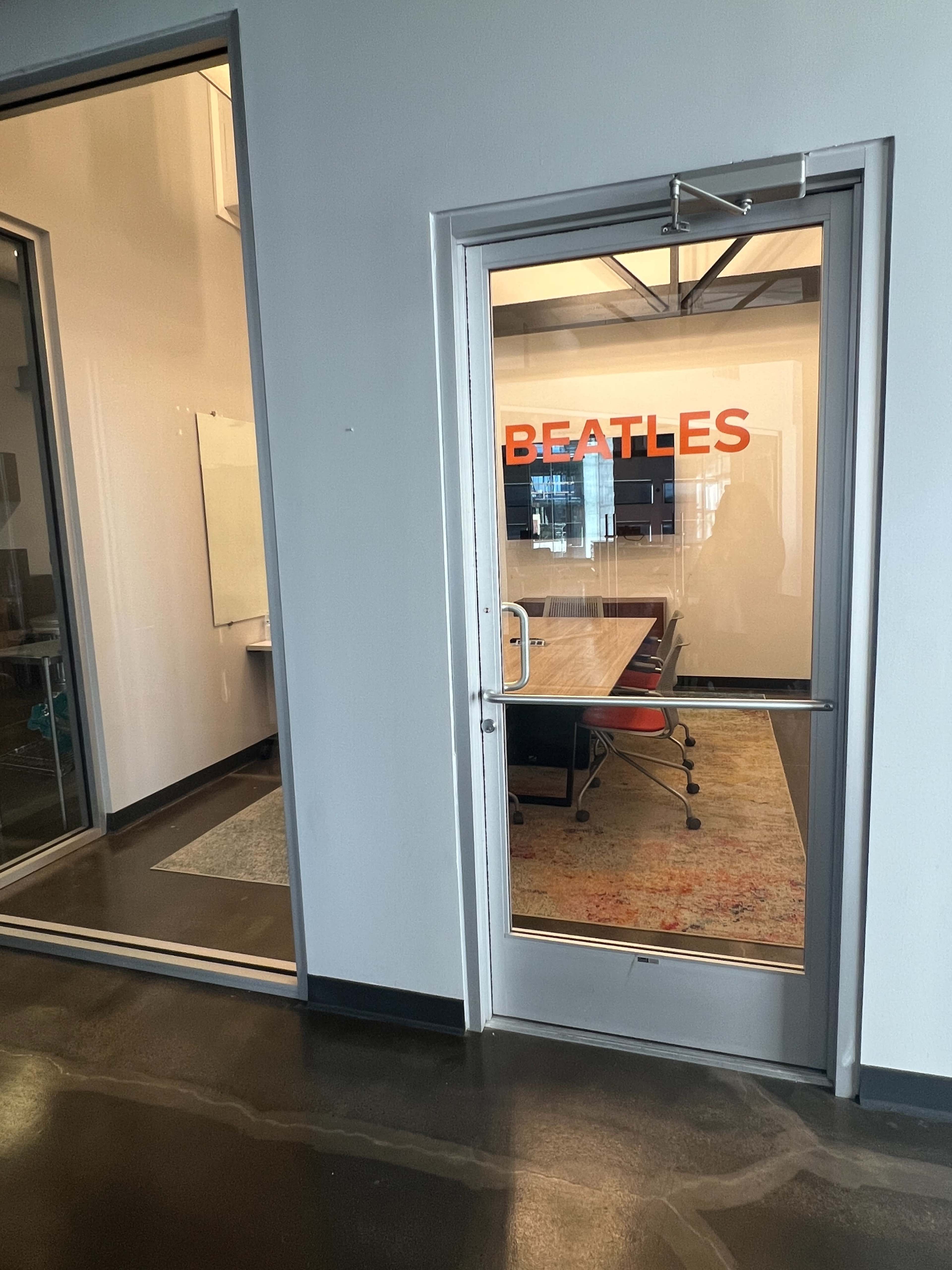 The image shows a glass door labeled "BEATLES" leading into a conference room with a visible table and chairs inside.