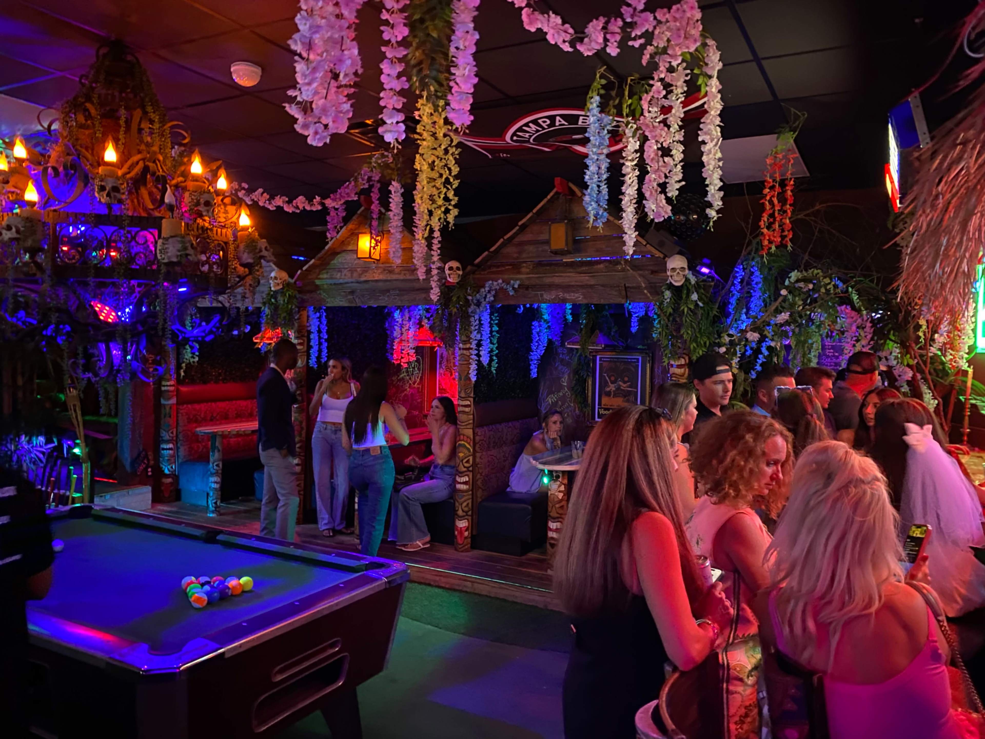 The image depicts a vibrant bar scene with patrons mingling around a pool table and a tiki-style seating area adorned with tropical decorations and colorful lighting.