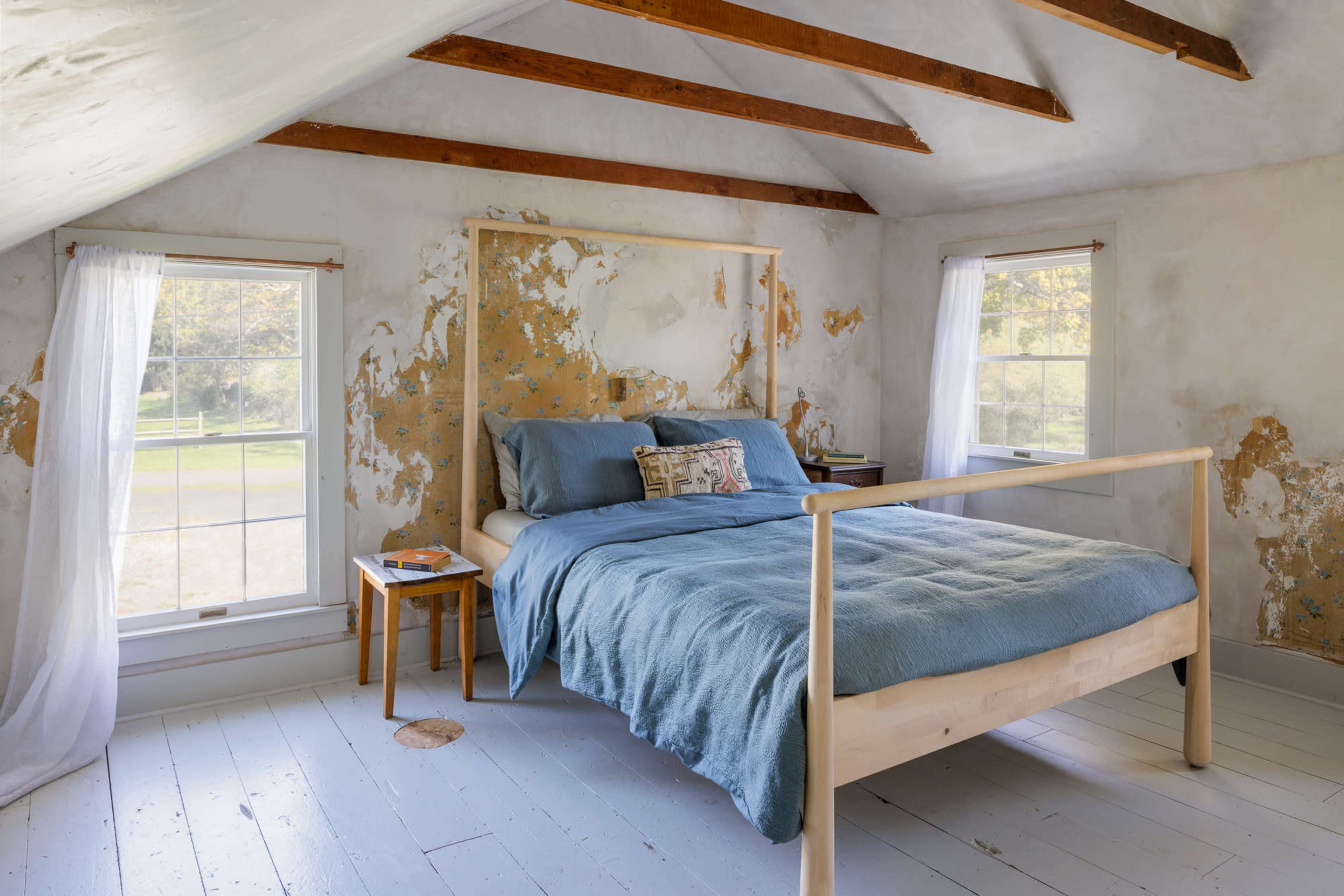The image shows a simple bedroom with a wooden bed, light blue bedding, and two windows revealing a view of the outdoors, all set against weathered walls.
