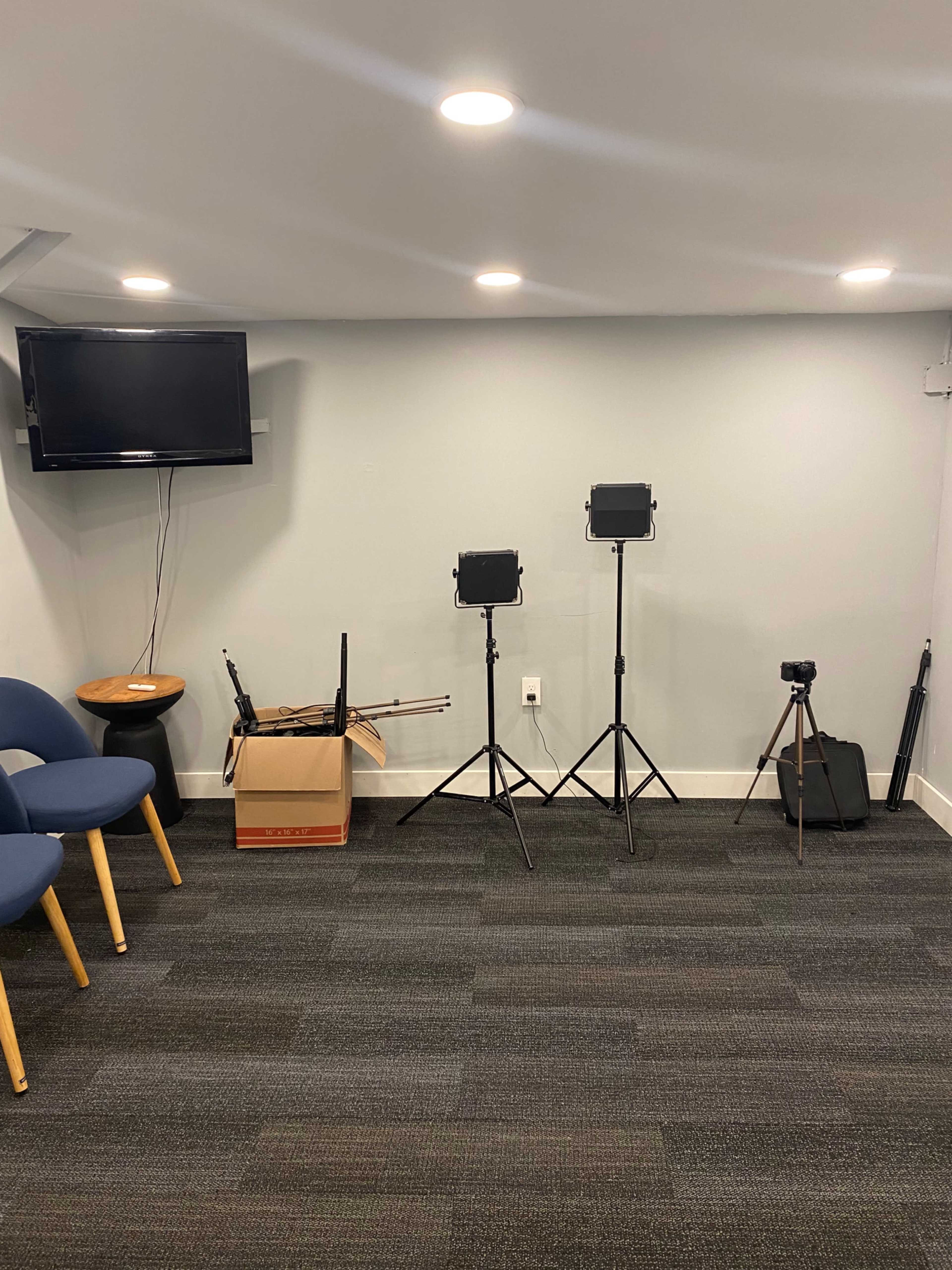 A small room with a television mounted on the wall, two lighting stands with softboxes, a box containing equipment, and a tripod, along with a few blue chairs arranged nearby.