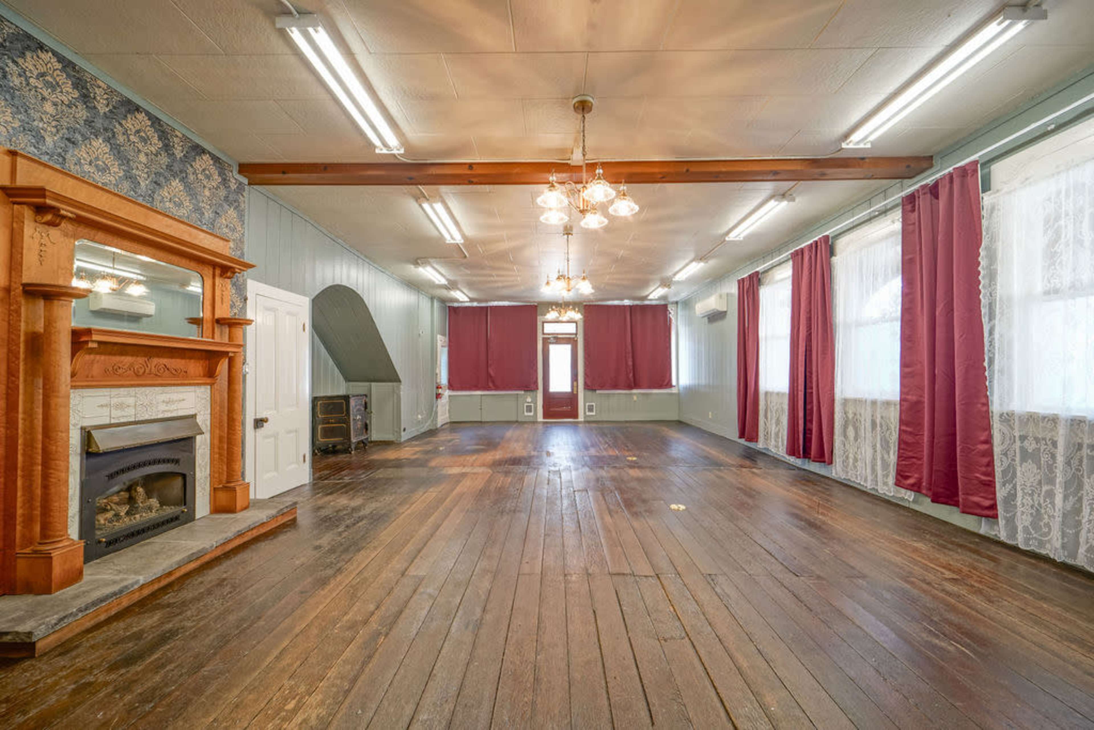 The image shows a spacious room with wooden floors, large windows covered by red curtains, and a decorative fireplace at one end.