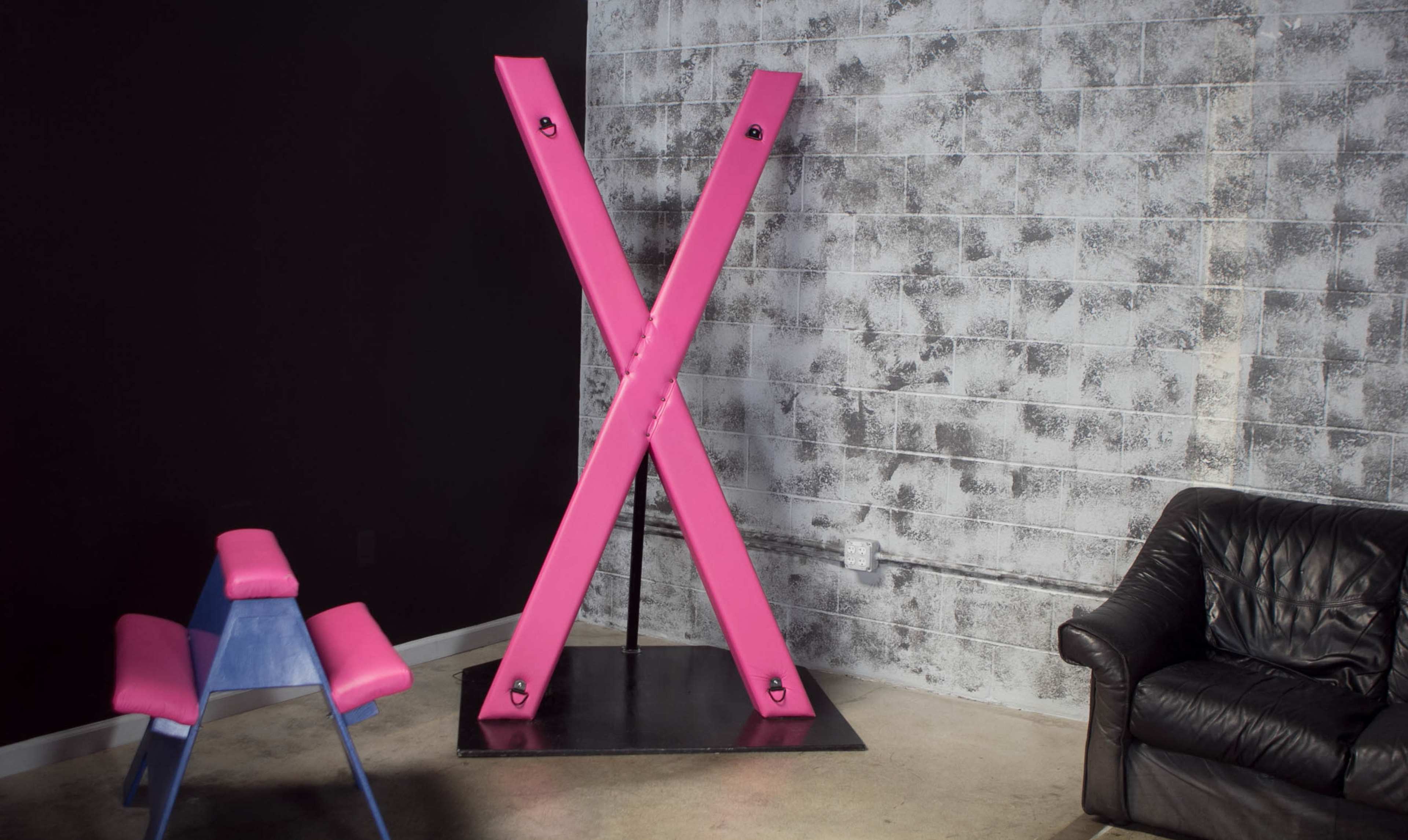 A large pink X-shaped structure against a textured gray wall, with a pink chair and a black sofa in the foreground.