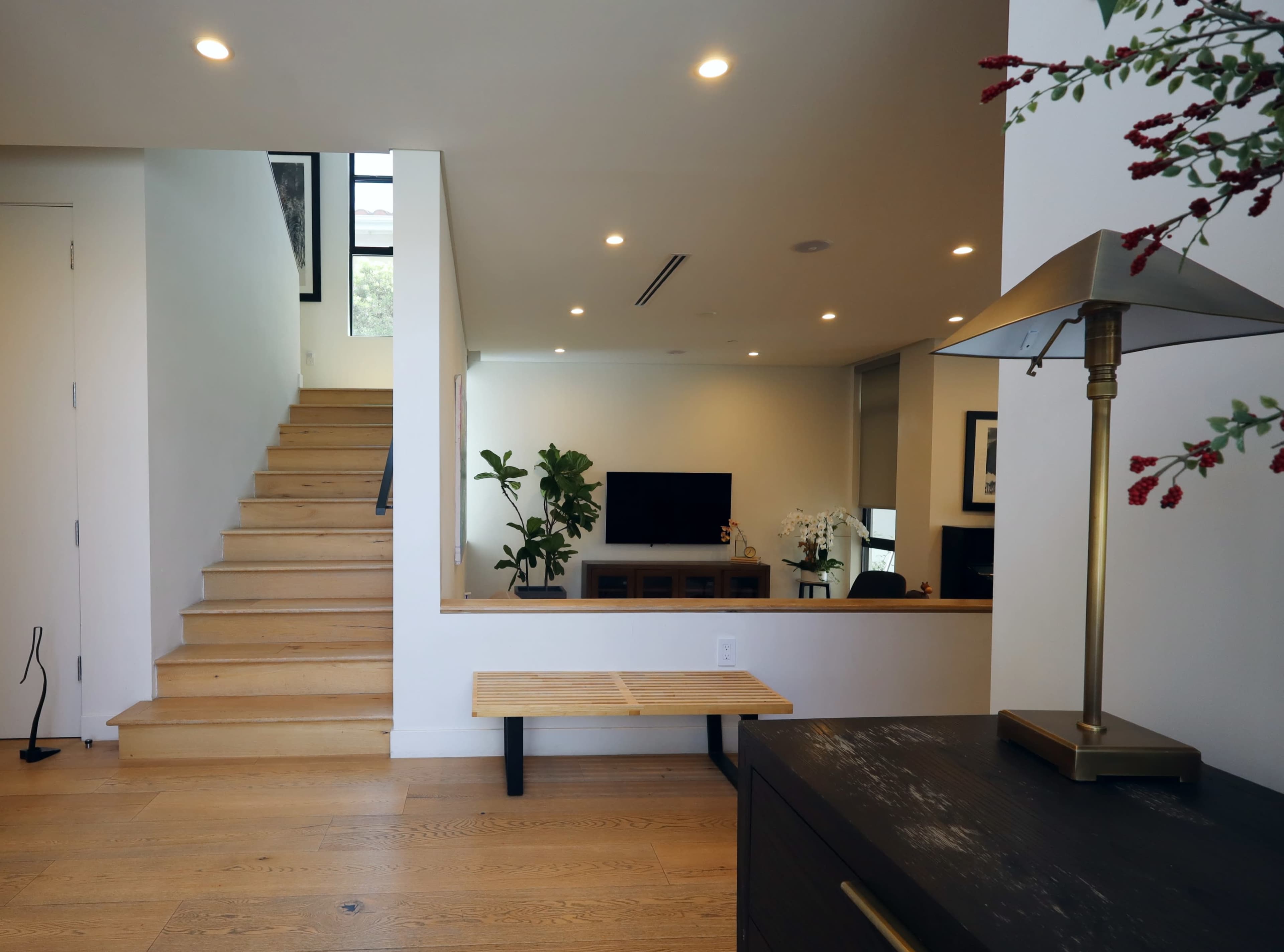 A wooden staircase leads to a bright living area featuring a television and plants, with a side table and lamp in the foreground.