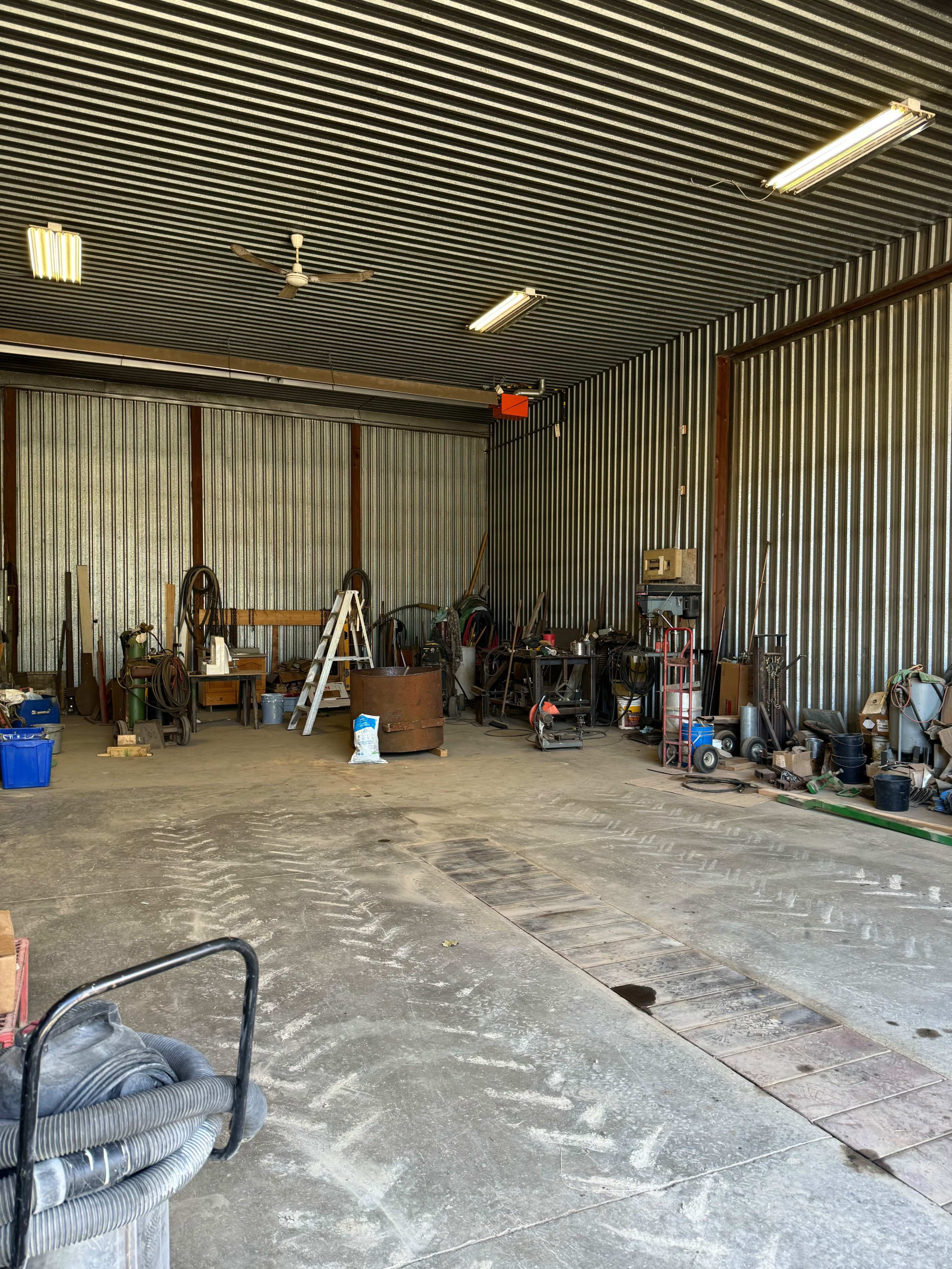 The image shows a spacious industrial workshop with various tools, equipment, and a ladder arranged along the walls, and a concrete floor marked with tire tracks.