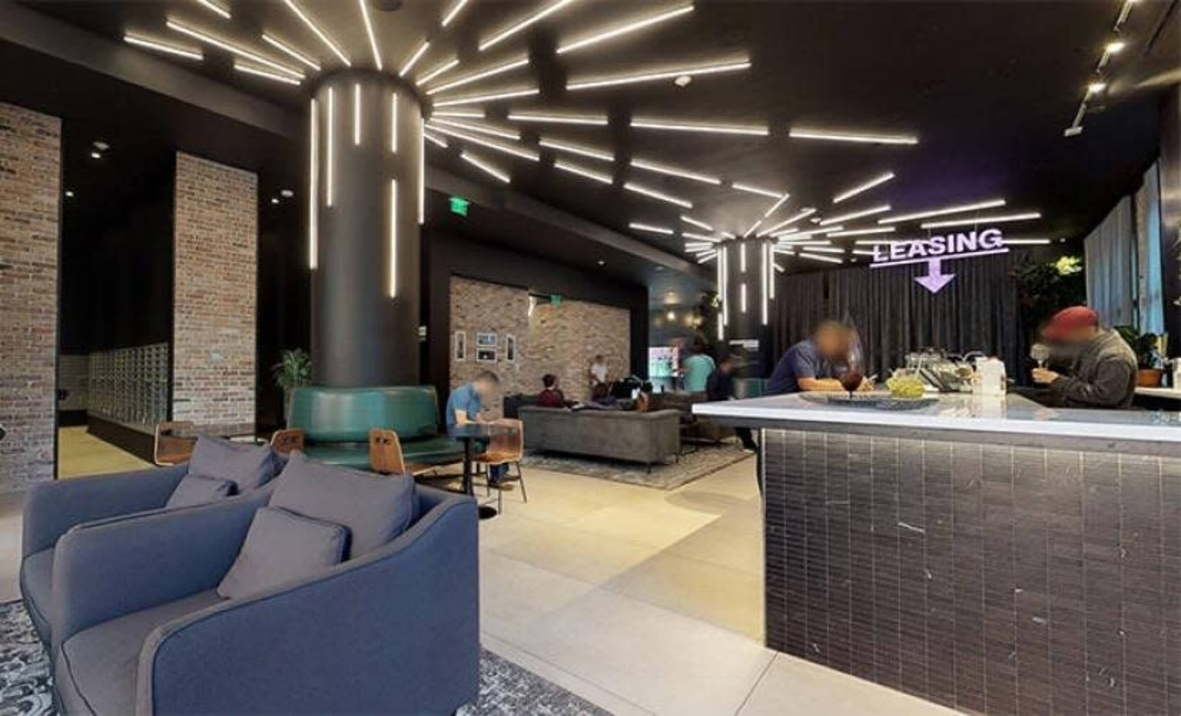The image shows a modern leasing office with stylish furniture, vibrant lighting, and a reception area featuring a leasing desk and seating arrangements.