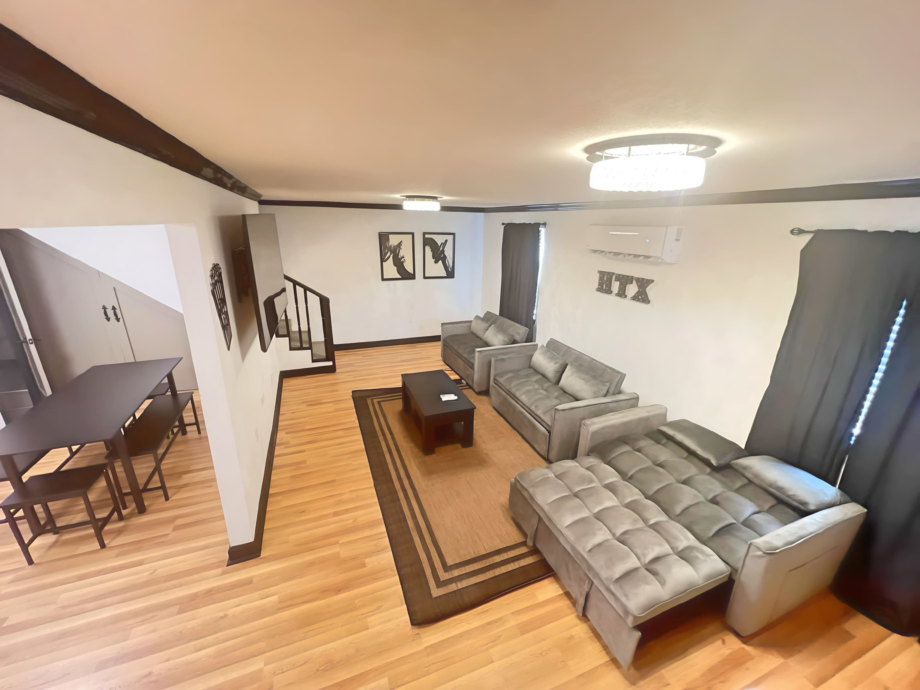 The image shows a modern living room with gray sofas, a coffee table, and a wooden floor, adjacent to a dining area with a table and chairs.
