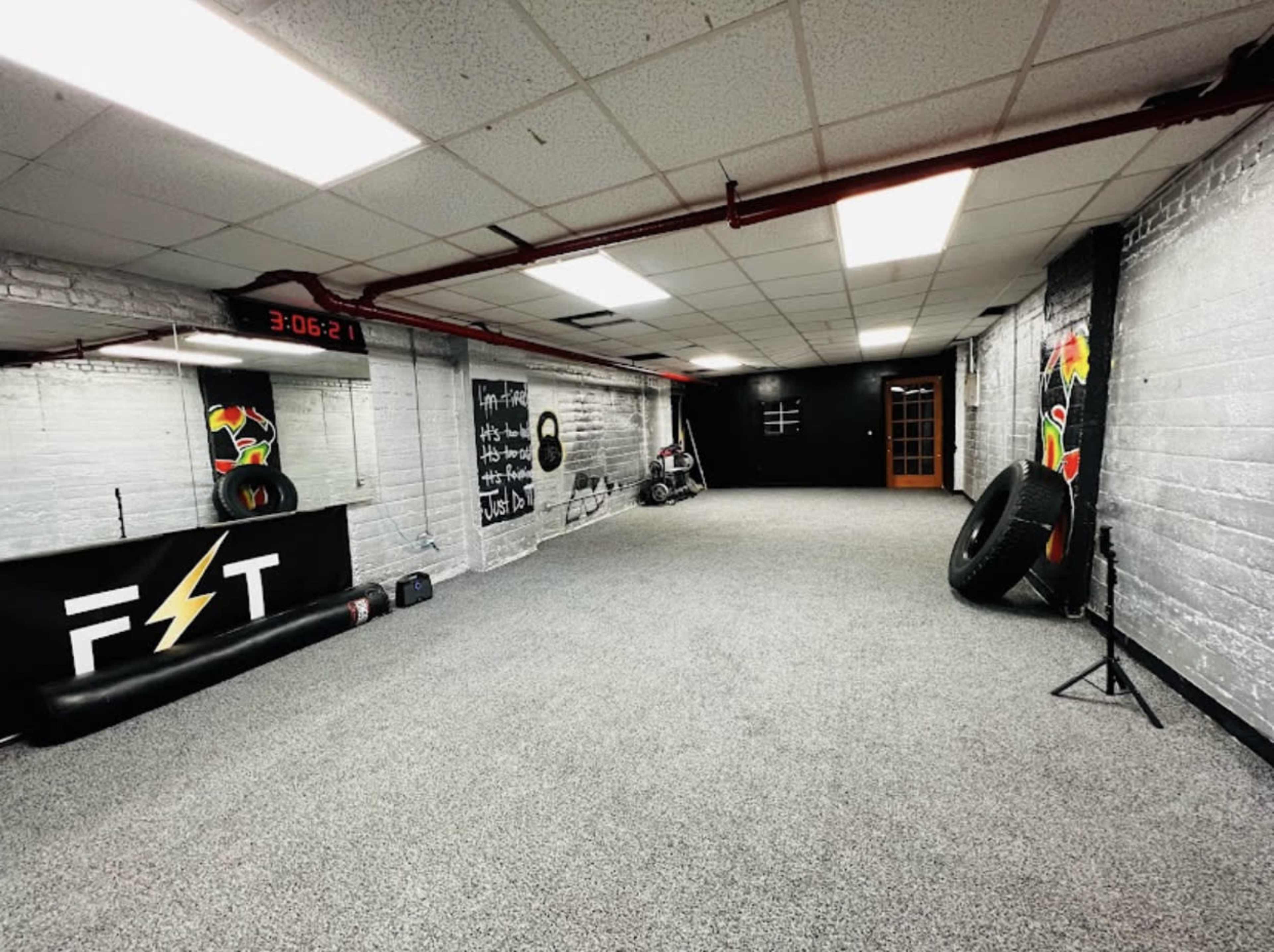An empty gym or training room with gray carpet, white brick walls, and fitness equipment positioned along the sides.